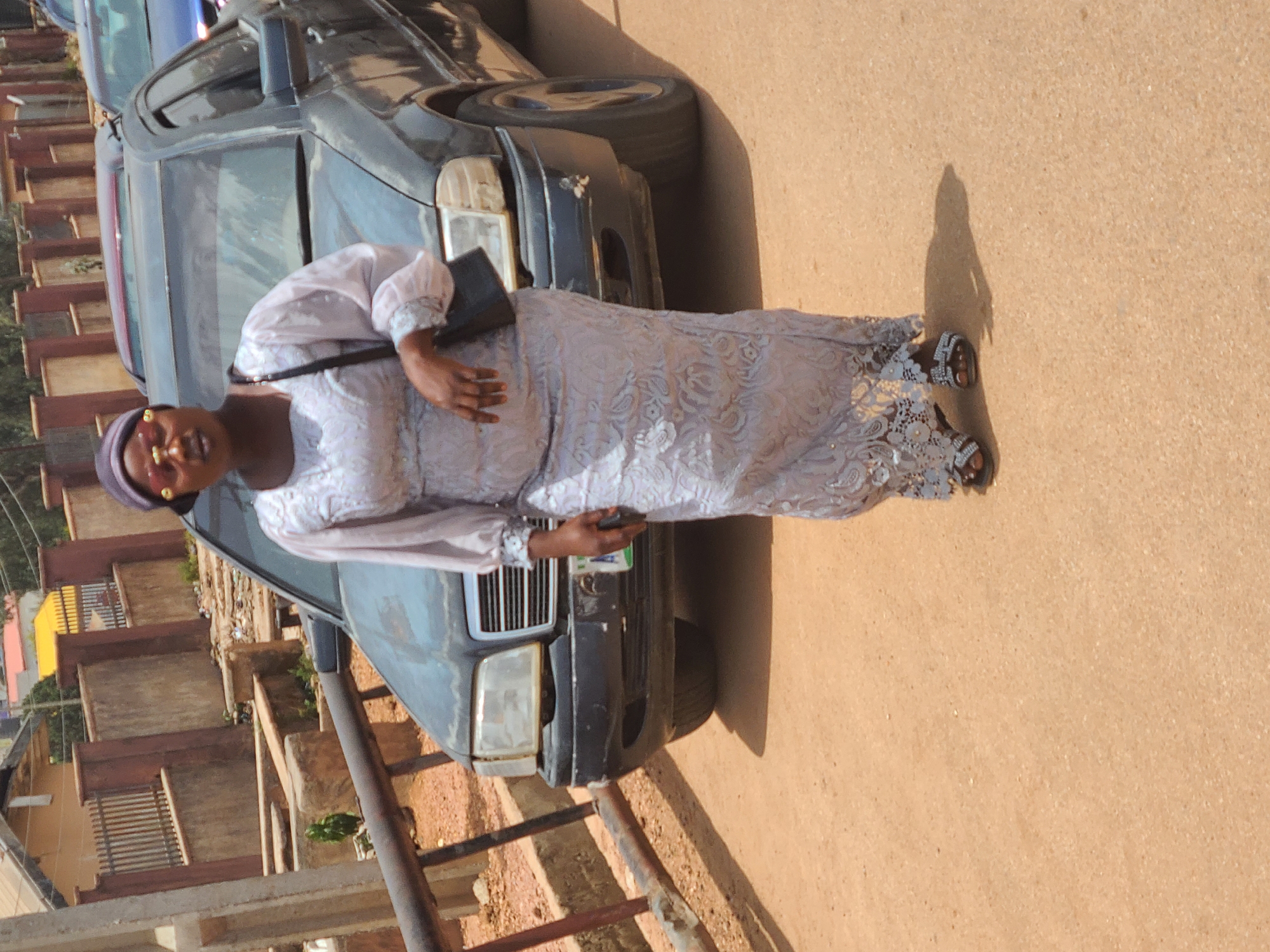 An adult African woman is standing on a paved road beside a dark colored sedan, possibly a Mercedes-Benz. She wears a long, pale dress with floral patterns and lace detailing, a dark head wrap, and sandals. She carries a small black purse on a strap over her shoulder and holds a mobile phone in her right hand. The background shows a partially obscured row of low buildings and greenery, typical of a residential or commercial area. The location is Jos, Nigeria. The weather appears bright, as indicated by the clear shadow cast by the woman on the road.