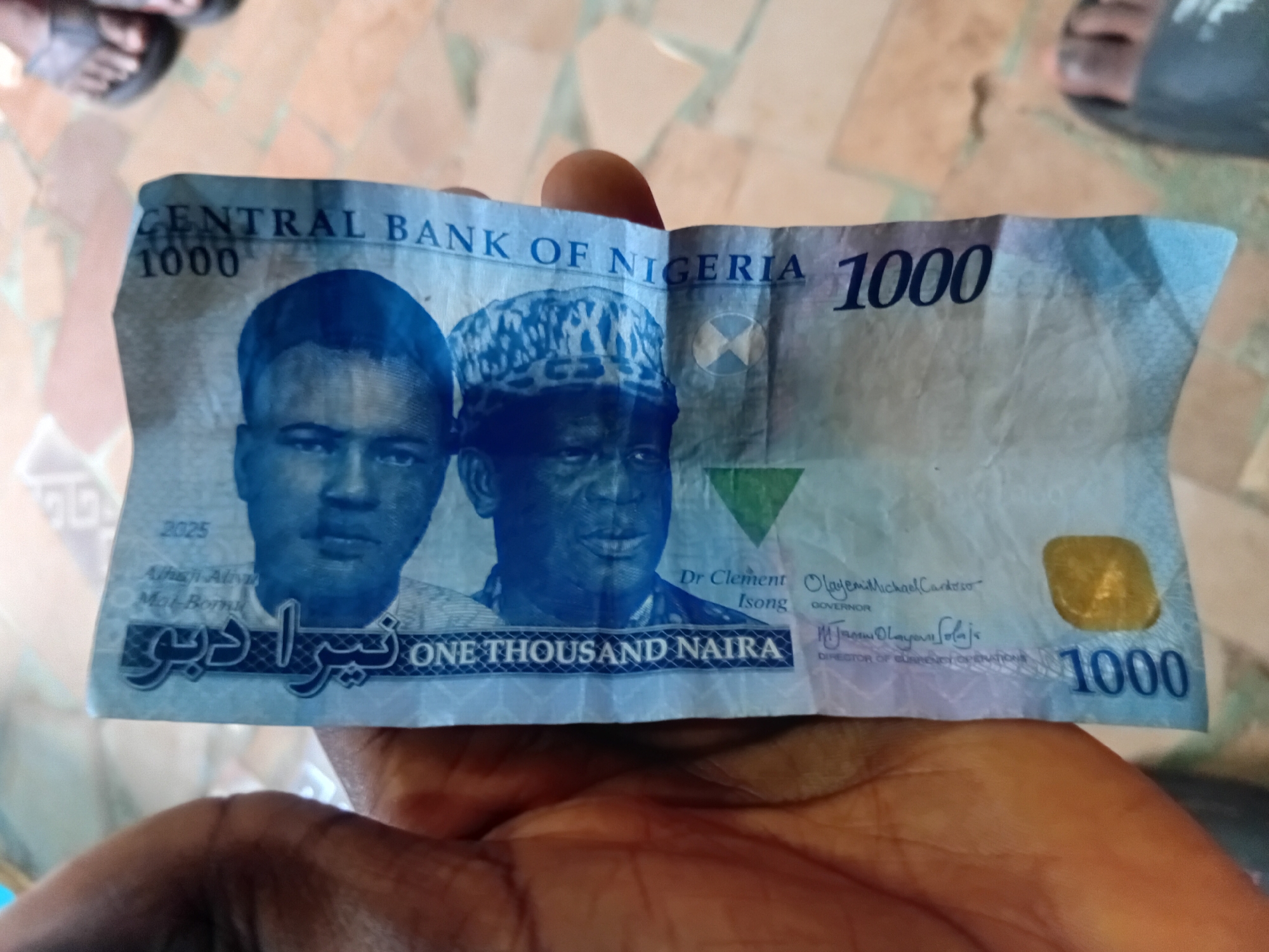 A person's hand is holding a Nigerian 1000 Naira banknote. The banknote is slightly creased and features portraits of two men, text in English and Arabic, and the number "1000" prominently displayed. The background is indistinct, suggesting an indoor or semi-outdoor setting. The image does not provide enough information to determine the specific location within Nkpor, Nigeria.