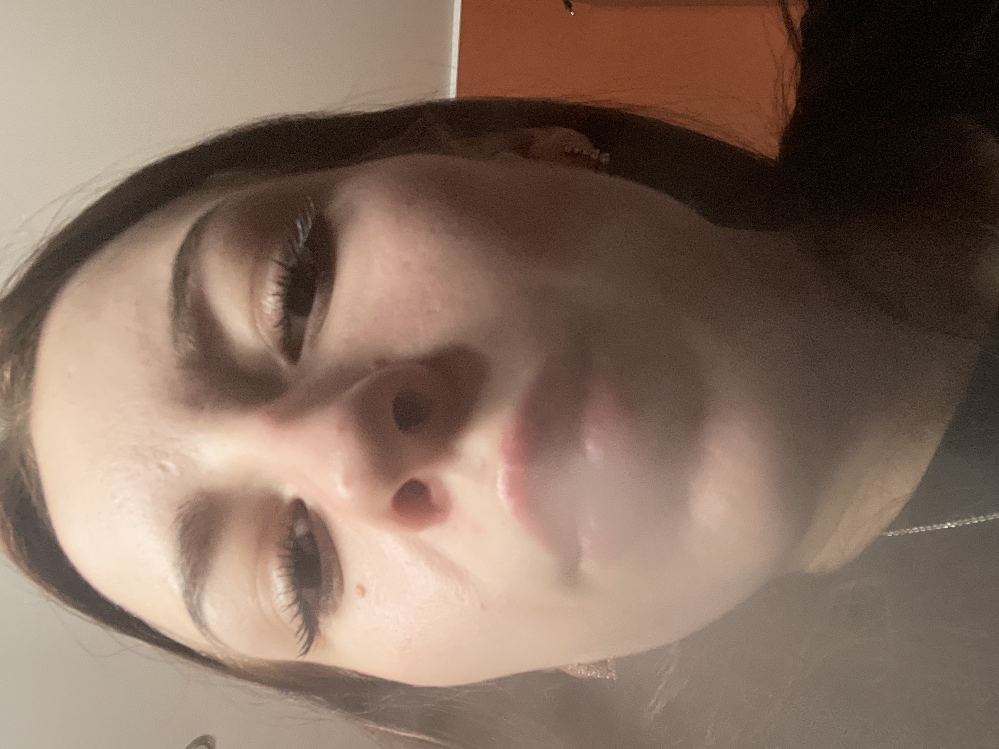 The image is a close-up, low-angle, selfie-style photograph of a young woman's face, taken from the chin upwards. The woman has light-colored skin, dark brown hair framing her face, and visible eyelashes. Her eyes are open, and her mouth is slightly parted, with a hazy, white substance obscuring her lower lip and chin, possibly smoke or steam. She is wearing small, sparkly earrings. The background is indistinct, appearing to be an indoor setting with a pale wall and an orange-colored surface visible to the right. There is a thin silver chain visible around her neck. The photograph appears to have been taken in the city of Медведево, Russia.