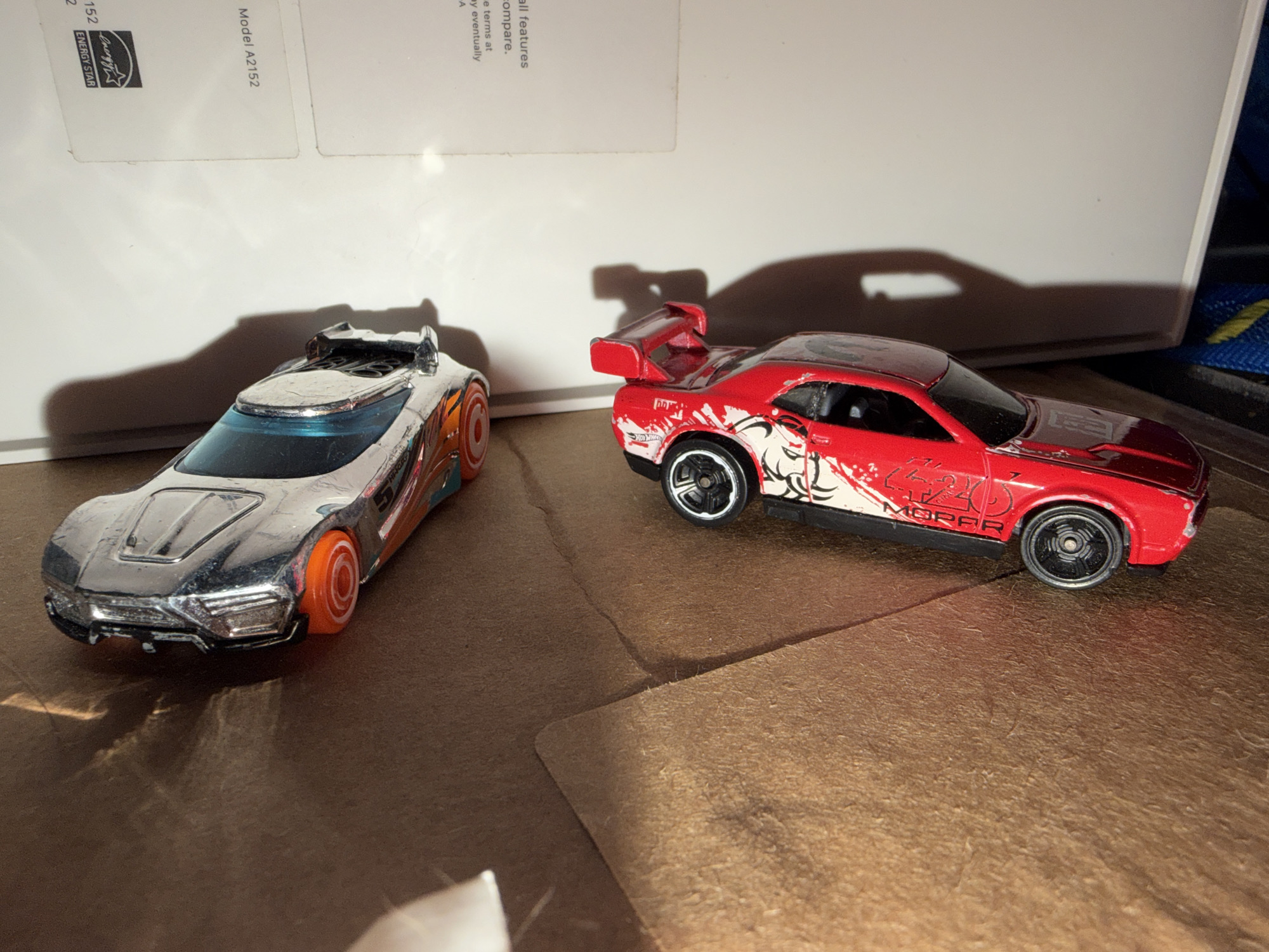 The image depicts two toy cars on a brown, textured surface, possibly cardboard. The car on the left is silver with orange accents and a blue windshield, featuring a futuristic design. The car on the right is red with white graphics, a large rear spoiler, and the word "Mopar" printed on its side. In the background, a white surface with text and logos is visible. The lighting creates shadows of the cars and other objects, suggesting an indoor setting. The scene appears to be a still life arrangement of toy vehicles. There is no discernible information about the location being Portland, United States of America.