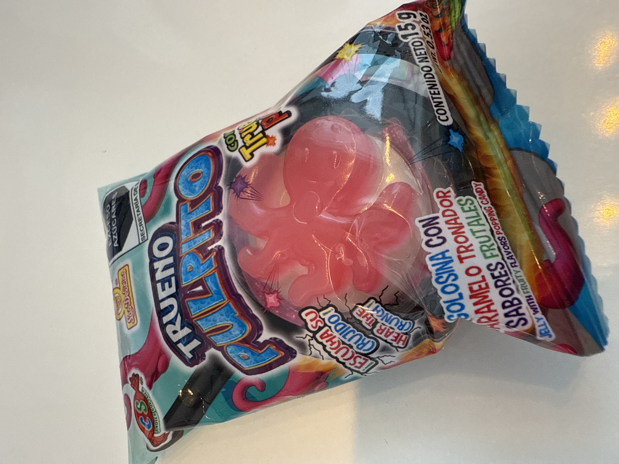 A close-up shot depicts a package of "Trueno Pulpo" candy. The packaging is predominantly clear with turquoise, pink, and black accents. An image of a pink octopus-shaped candy is visible within the package. Text on the package includes phrases like "Escucha su crujido!" ("Hear the crunch!"), "Solosina con caramelo tronador sabores frutales" ("Jelly with fruity flavors popping candy"), and "Contenido neto 15 g" ("Net content 15 g"). Additional text includes "Exceso Azúcares" ("Excess Sugars"). The brand logo "C&S Candies Supplies Sas Delicias" is also present. The background is a plain white surface. There is no indication of this image being taken in the city of Puebla, Mexico.