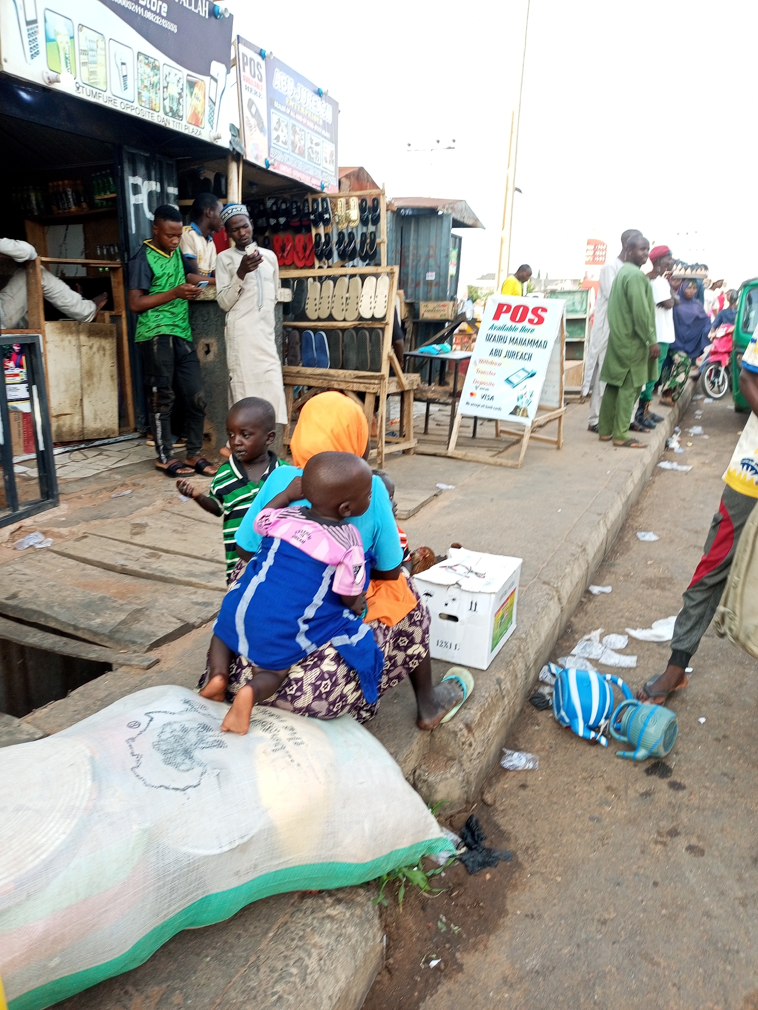 An outdoor street scene shows a gathering of people, including adults and children, in what appears to be a market or commercial area. In the foreground, a woman wearing an orange headscarf and a patterned skirt is holding two young children. One child is dressed in a blue and pink outfit, and the other wears a green and black striped shirt. They are sitting on a large, light-colored bag that is placed on a wooden surface.
To the left, a young boy in a green t-shirt and black pants is looking at a mobile phone. He stands near a stall with a sign advertising mobile phones and accessories. Another man in a white robe and cap is also looking at a phone. In the background, other people are visible, some walking and others standing. A sign for "POS" with information about money transfer services is displayed prominently. Several stalls are set up with goods, including footwear and other merchandise. The ground is paved with concrete and some areas are wooden. The overall environment suggests a busy urban marketplace.