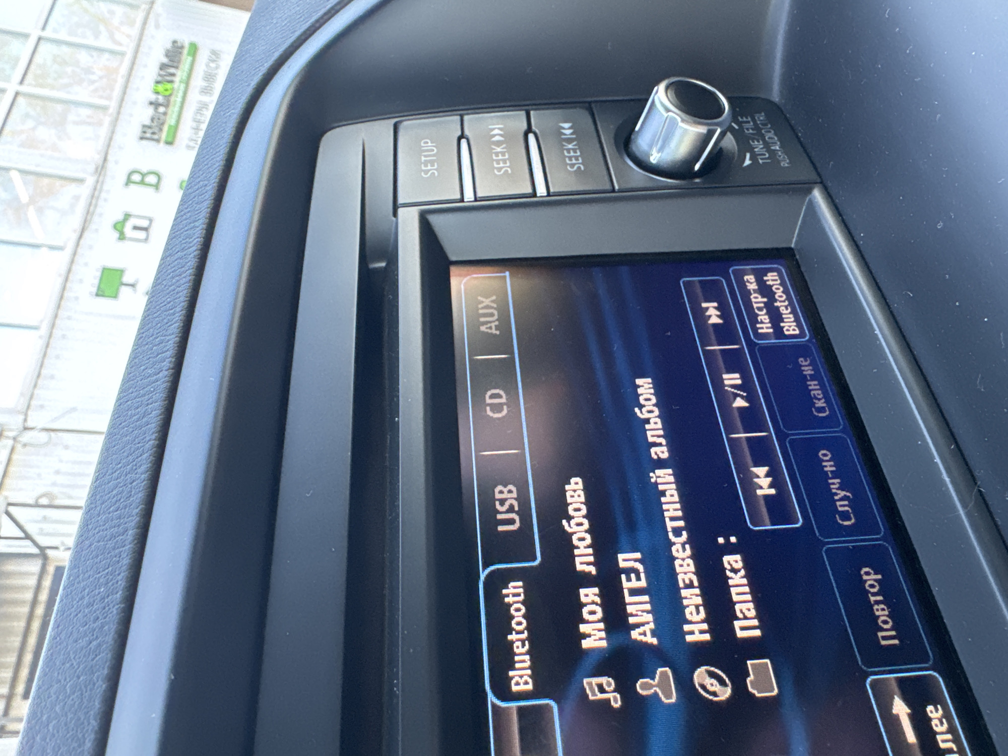Here is a description of the image:
The image shows the interior of a car, specifically the car's infotainment system display. The system is set to play music via Bluetooth, with the song "Моя любовь" (My Love) by АИГЕЛ (AIJEL) selected. The screen also displays the album and folder details, which are in Russian. Buttons for setup, seek forward, and seek backward are visible above the screen. A volume control knob is located on the right. In the background, out the window, there is a sign that says "Black & White" and also includes what appears to be a street address in Russian. The scene is likely inside a car in Chaikovsky, Russia, given the presence of Russian text and the specific street signage.