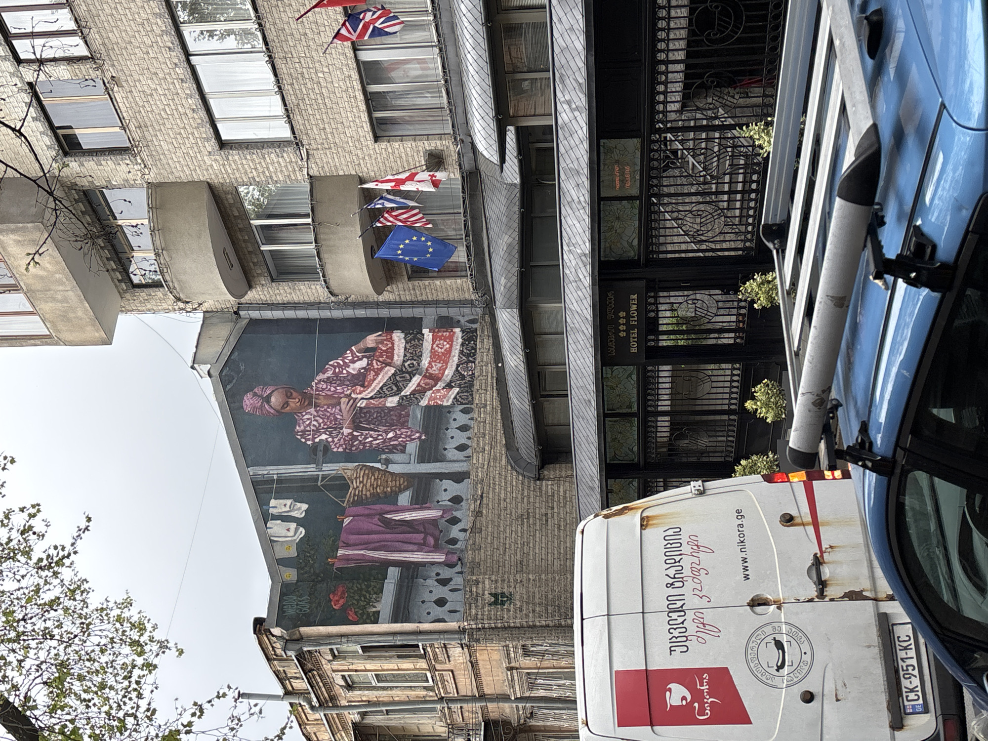 The image captures a street scene in Tbilisi, Georgia. A mural depicting a woman in traditional clothing and a clothesline is painted on the side of a brick building. Above the mural are several flags, including the United Kingdom, Georgian, American, and European Union flags. Adjacent to the building with the mural is a gated entrance with a sign that reads "Hotel Flower" in both English and Georgian. The foreground features a white van and a blue car, obscuring part of the view. The van has Georgian writing and the website "www.nikora.ge" printed on it, as well as license plate "CK-951-KC" which indicates that it is registered in Georgia. Part of a tree is visible on the left side of the image. The angle of the shot suggests the photograph was taken from a low vantage point, looking upwards.