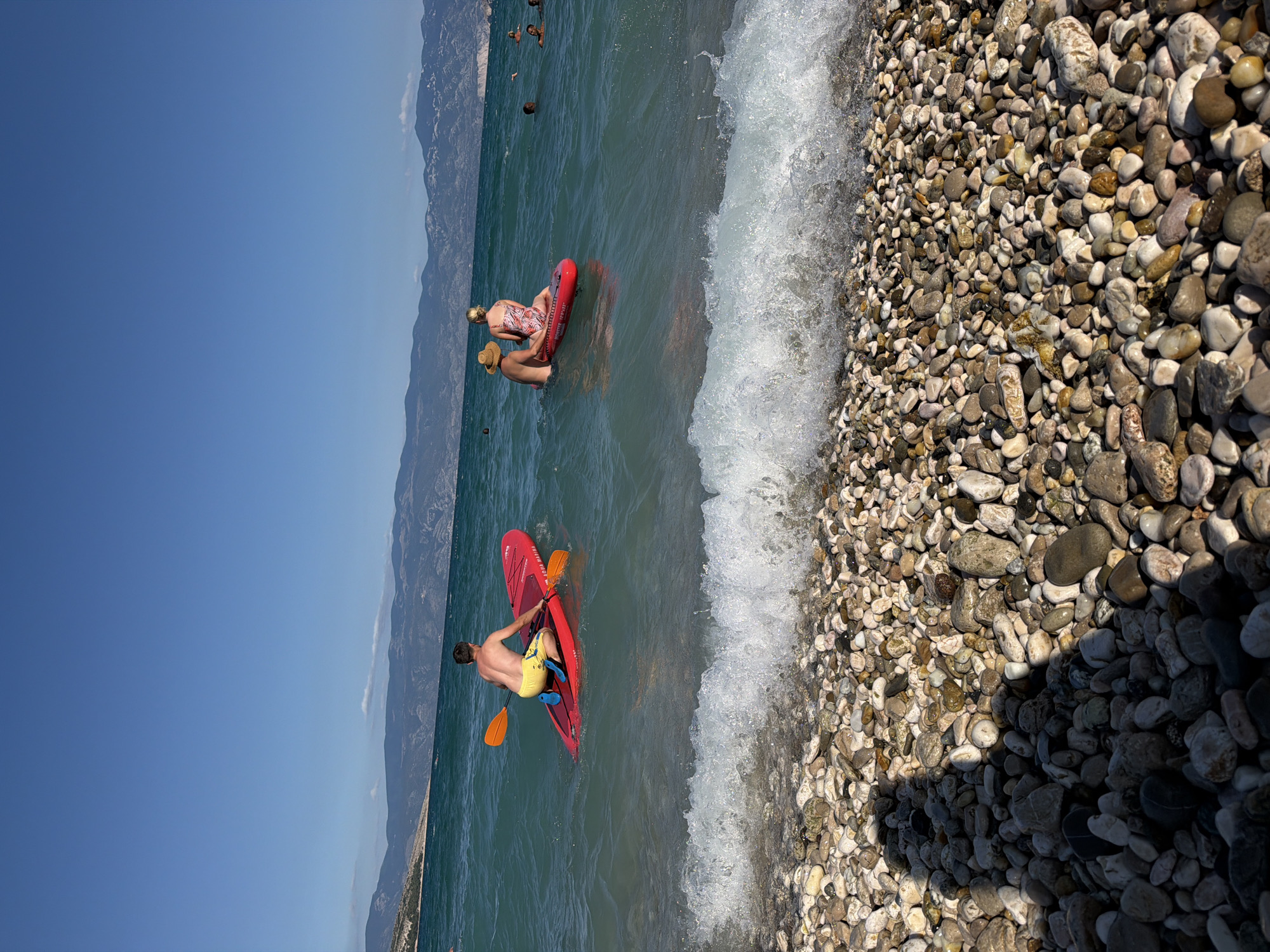 The image depicts a sunny day at a pebble beach in Baška, Općina Baška, Croatia. In the foreground, the beach is covered in smooth, multi-colored pebbles, with a gentle wave breaking at the waterline. In the middle ground, two individuals are on red stand-up paddleboards in the turquoise water. One person, wearing yellow shorts, is paddling a board with an orange paddle. Another person, wearing a patterned swimsuit and a straw hat, is sitting on a board with someone else behind them, also in the water. In the background, there are hazy mountains under a clear blue sky, suggesting a coastal mountain range. Several other people are visible swimming or floating in the water further out.