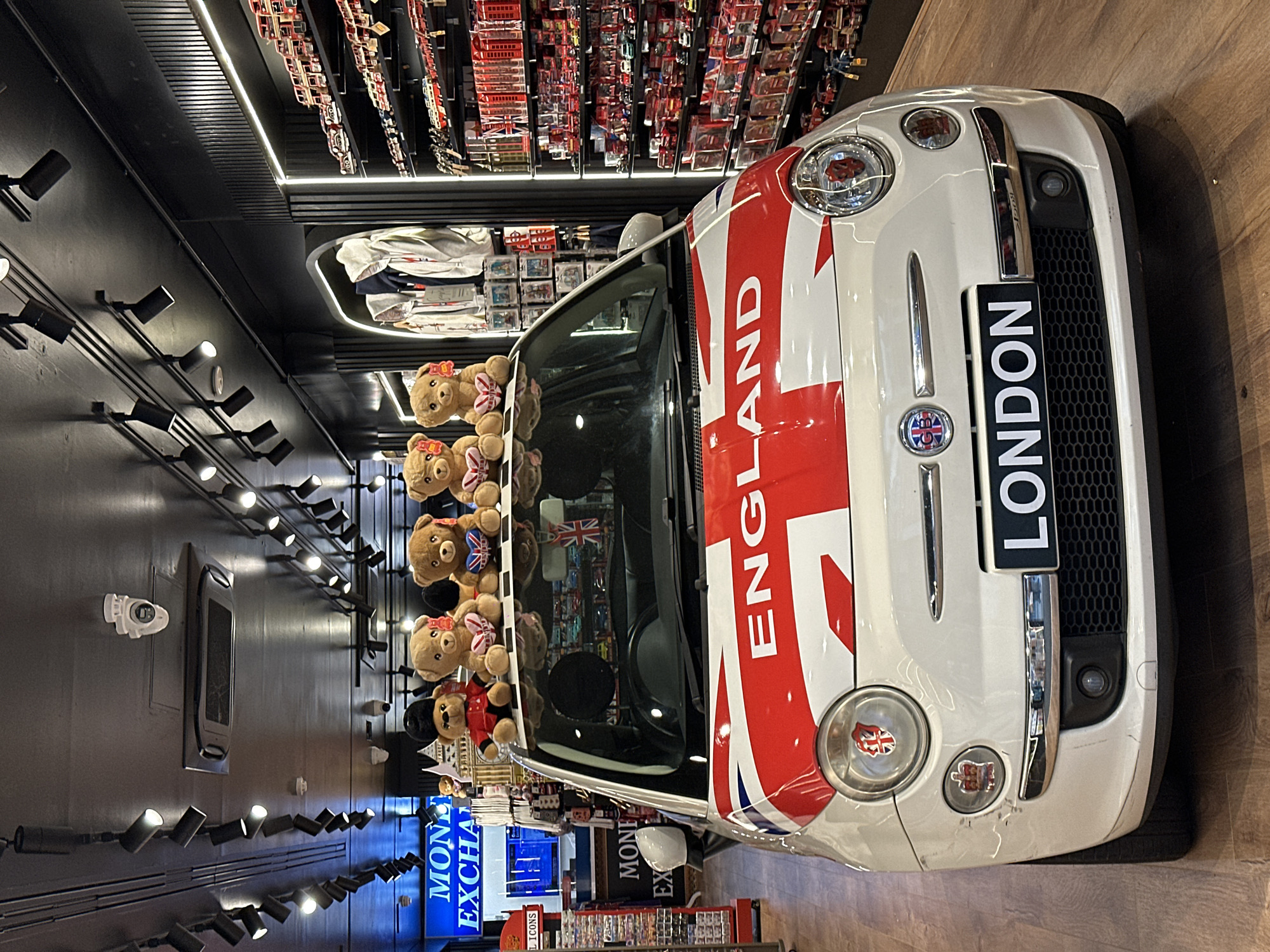 A car with an "ENGLAND" theme, featuring the colors red, white, and blue, is parked in a store in London. The car has a license plate that reads "LONDON". There are teddy bears on the roof of the car. The store sells London-themed merchandise, including items related to the British flag and the United Kingdom. There is a "MONEY EXCHANGE" sign in the store. The wall has a series of lights.