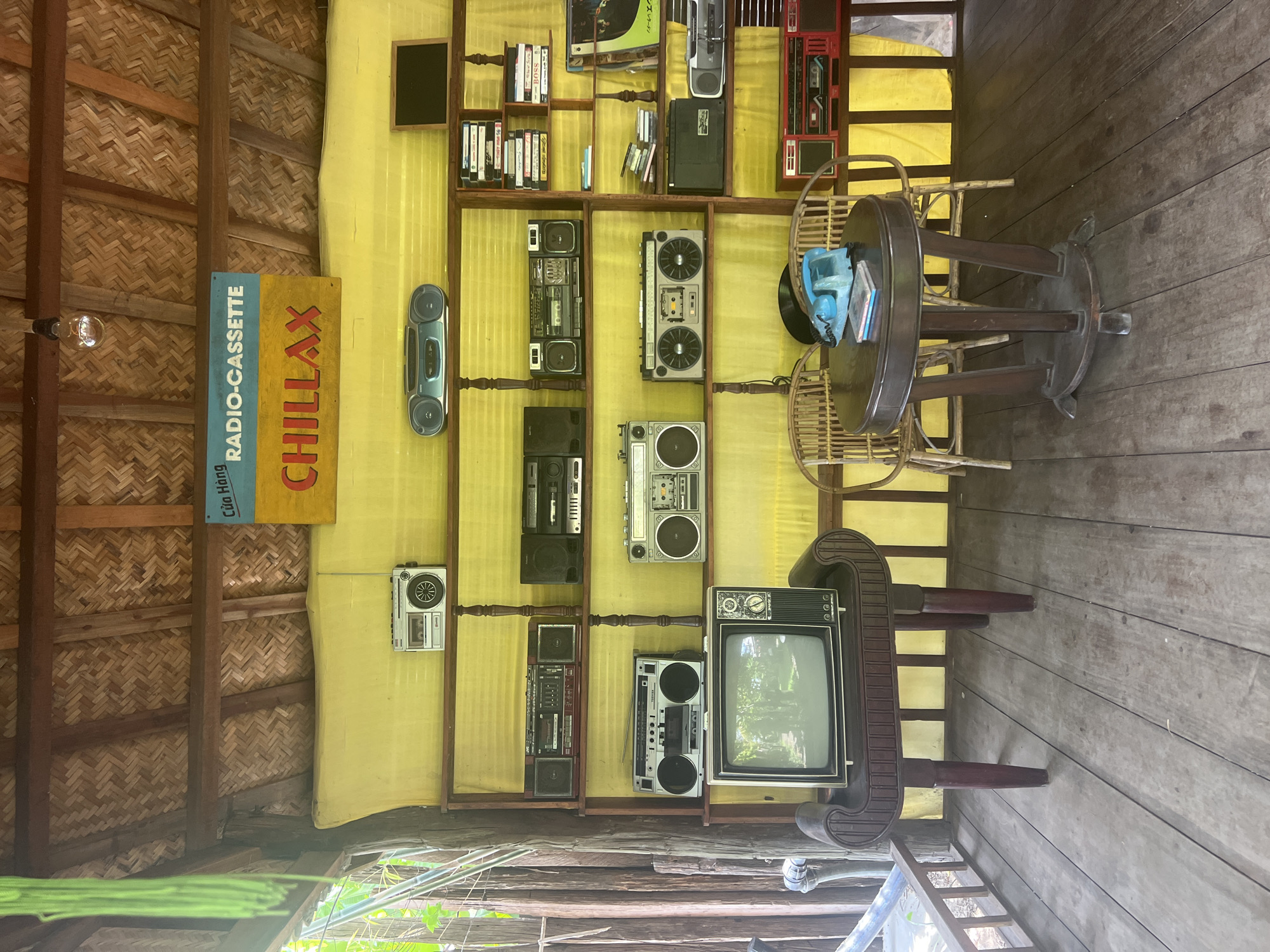This image shows a display of vintage audio equipment and a television in an open-air structure with a thatched roof and wooden floor, likely a shop or cafe in Hoi An, Vietnam. The wall behind the display is covered with yellow fabric. On a shelf, there are several boomboxes and a smaller silver radio. Below this, a vintage television sits on an ornate wooden stand. To the right, more shelves hold various electronic devices, including cassette tapes and what appear to be records. In the foreground, a round wooden table with two wicker chairs is set up, with a blue rotary telephone and some books on the table. A sign above the display reads "Cửa Hàng RADIO-CASSETTE CHILLAX." The overall impression is one of a retro-themed establishment.