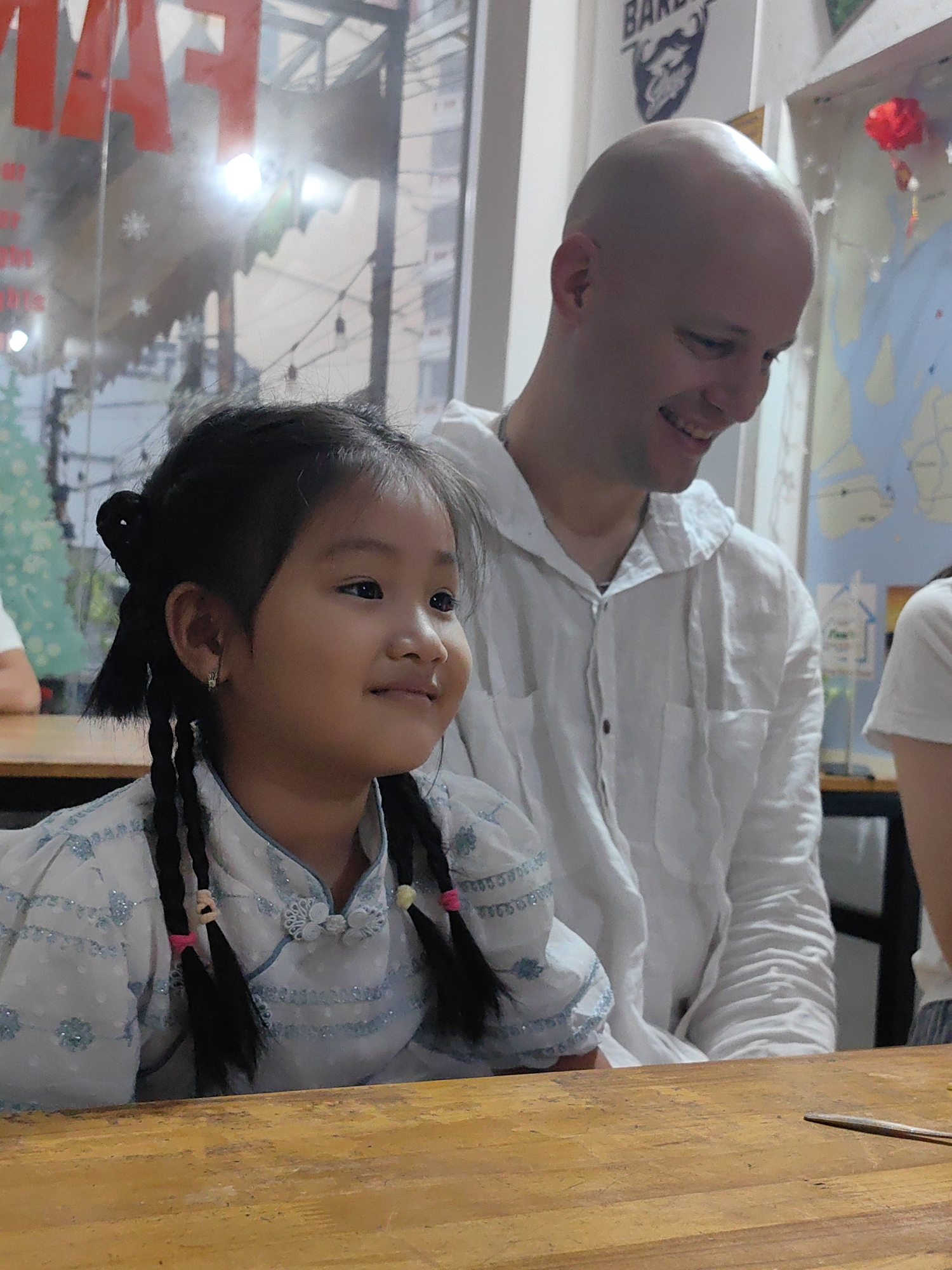 A young girl with braided hair and a white dress sits at a wooden table next to a bald man in a white shirt. Both are looking to the right. The background includes a window with reflective text, a Barber Cafe sign, and a map. The location is likely Bai Chay Ward, Vietnam, based on visual cues from the scene.