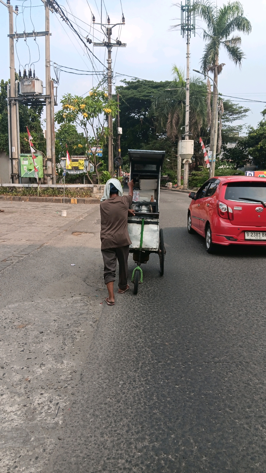 An Indonesian man wearing a brown t-shirt, brown pants, and a white head covering is pushing a food cart on a paved road. The food cart appears to be made of metal and has two large wheels in the back and one smaller wheel in the front. The cart is covered with a black tarp or lid. In the background, there are electrical poles with many wires, a cell tower, lush green trees, and a red car with the license plate B 2381 BK. The scene appears to be on the side of a road in Depok, Indonesia, likely during daylight hours given the lighting.