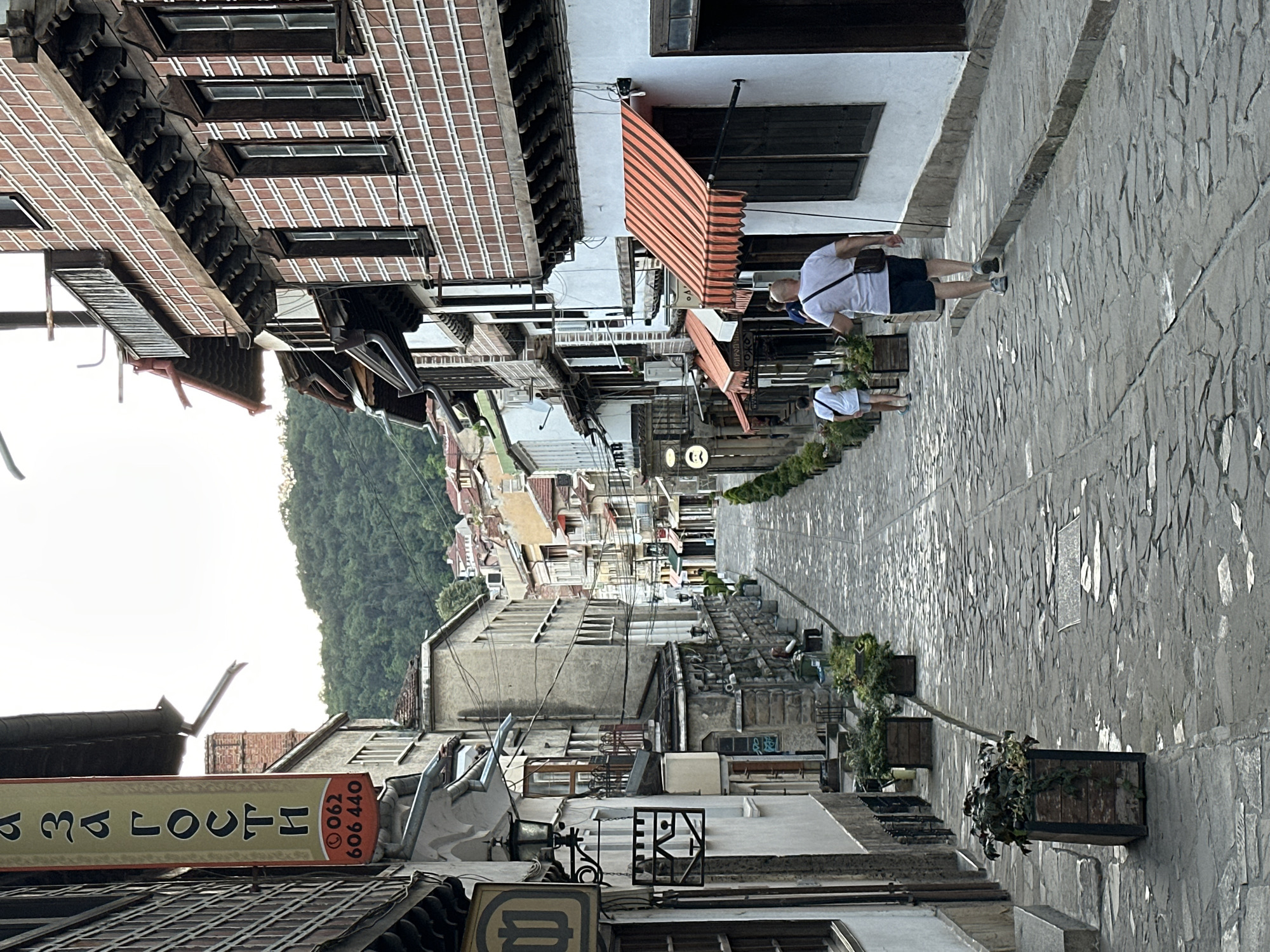 The image captures a view of a cobblestone street in Veliko Tarnovo, Bulgaria, looking uphill towards forested hills. On the left side of the street, a building features a prominent vertical sign with Cyrillic text and a phone number, along with decorative architectural elements. Further up the street on the left, more buildings with traditional architecture are visible. On the right side of the street, a building displays a patterned brick facade with numerous windows and wooden balconies. A striped orange awning extends over a storefront. Two individuals are walking uphill on the right side of the street, one in the foreground wearing a white t-shirt and dark shorts, carrying a bag, and another person ahead of them in a white shirt and shorts. The street is lined with potted plants. The overall scene depicts a charming, historic street in the city.