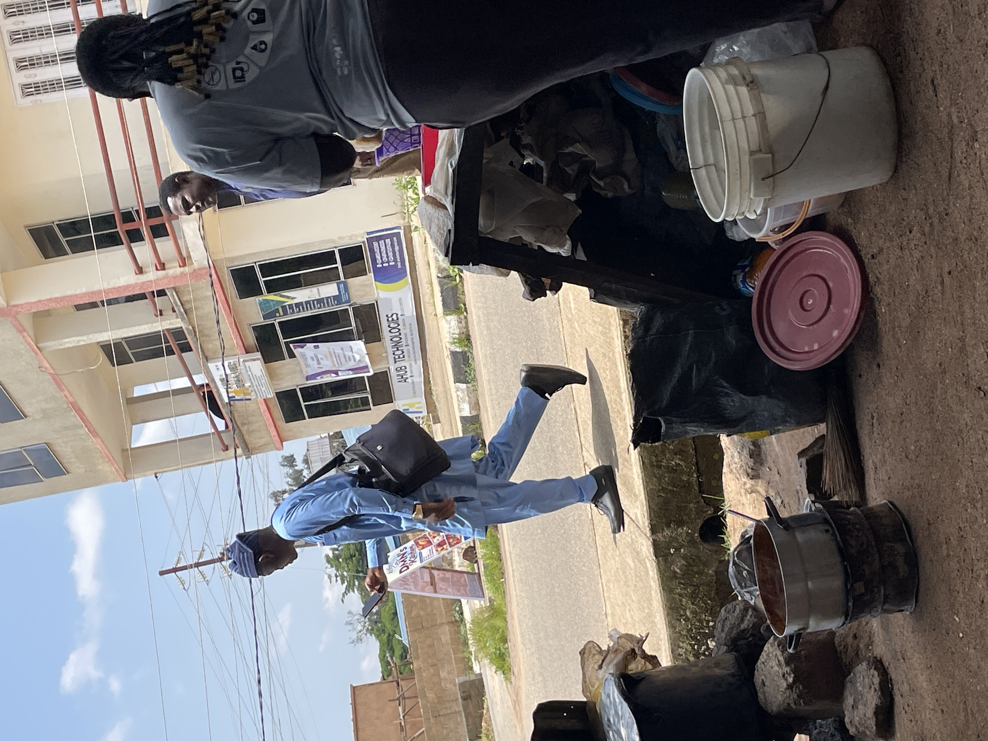 A street scene in Jos, Nigeria, captures a man in a blue outfit walking while looking at his phone, a black bag slung over his shoulder. He is walking on a sidewalk in front of a building. In the foreground, a cooking pot is placed on a makeshift stove, surrounded by other assorted objects, including buckets and plastic bags. The background shows a multi-story building with balconies and windows, suggesting an urban setting. The sky is clear with some clouds.