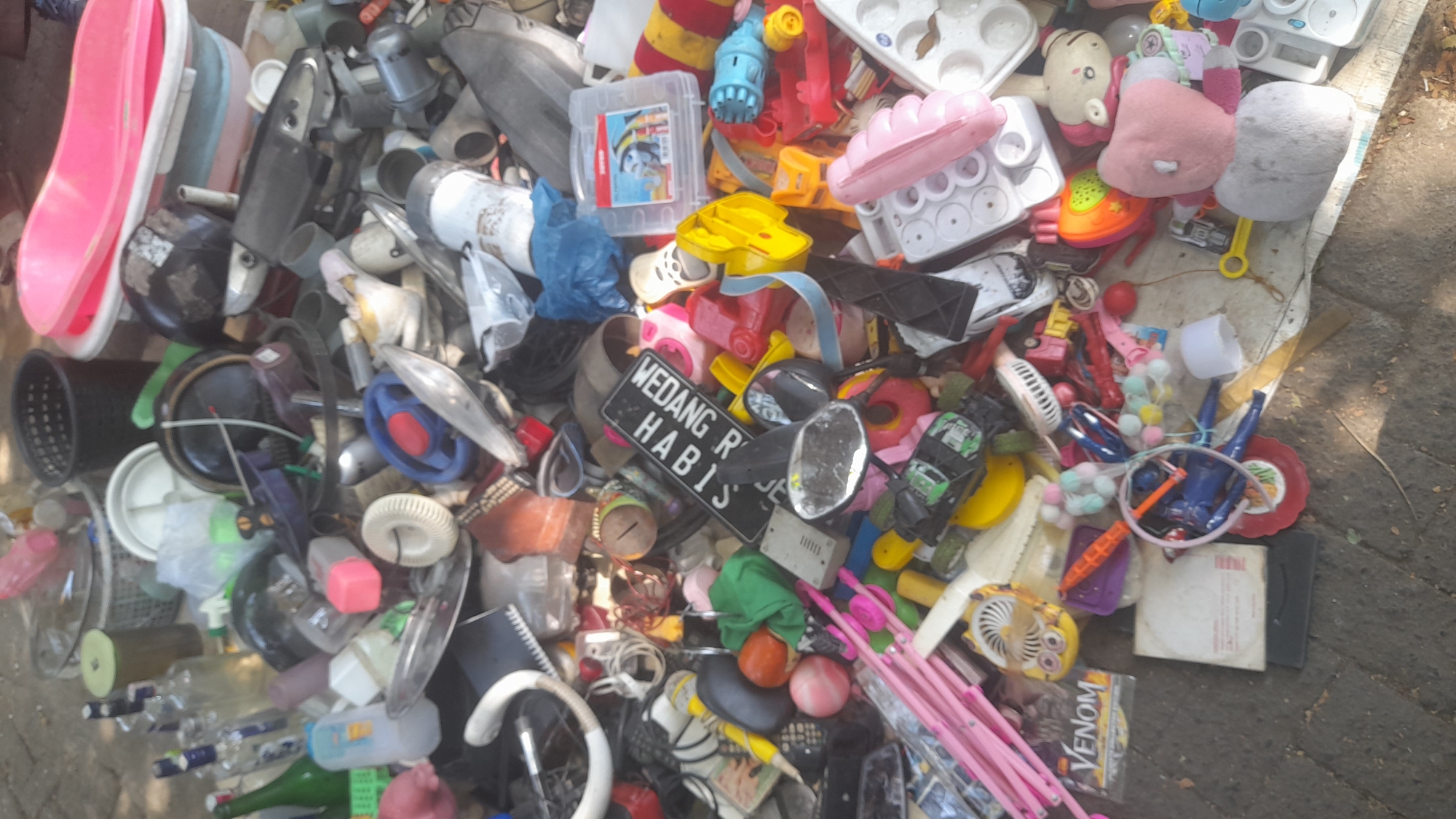 The image depicts a jumbled collection of assorted items, appearing to be discarded or for sale, spread on a paved ground surface. The scene is outdoors, likely in an urban environment, given the paving stones and the presence of various manufactured goods. Among the items are numerous toys, including small cars, dolls, and colorful plastic objects. There are also household items like plastic containers, a pink bathtub, a black basket, and what appears to be a white washbasin. Some electronics or their parts are visible, such as a white fan and a small black rectangular object with a white border that could be a broken screen. A sign with Indonesian text, "WEDANG HABIS," is prominently displayed. Several bottles, some appearing to contain liquids and others empty, are also present, along with various plastic bags and wrappers. The overall impression is one of disarray and potential waste or a makeshift market stall. The location is identified as Pekalongan, Indonesia, suggesting a possible scene from a local market, roadside vendor, or a collection point for recyclable materials. No people are clearly visible in the frame.