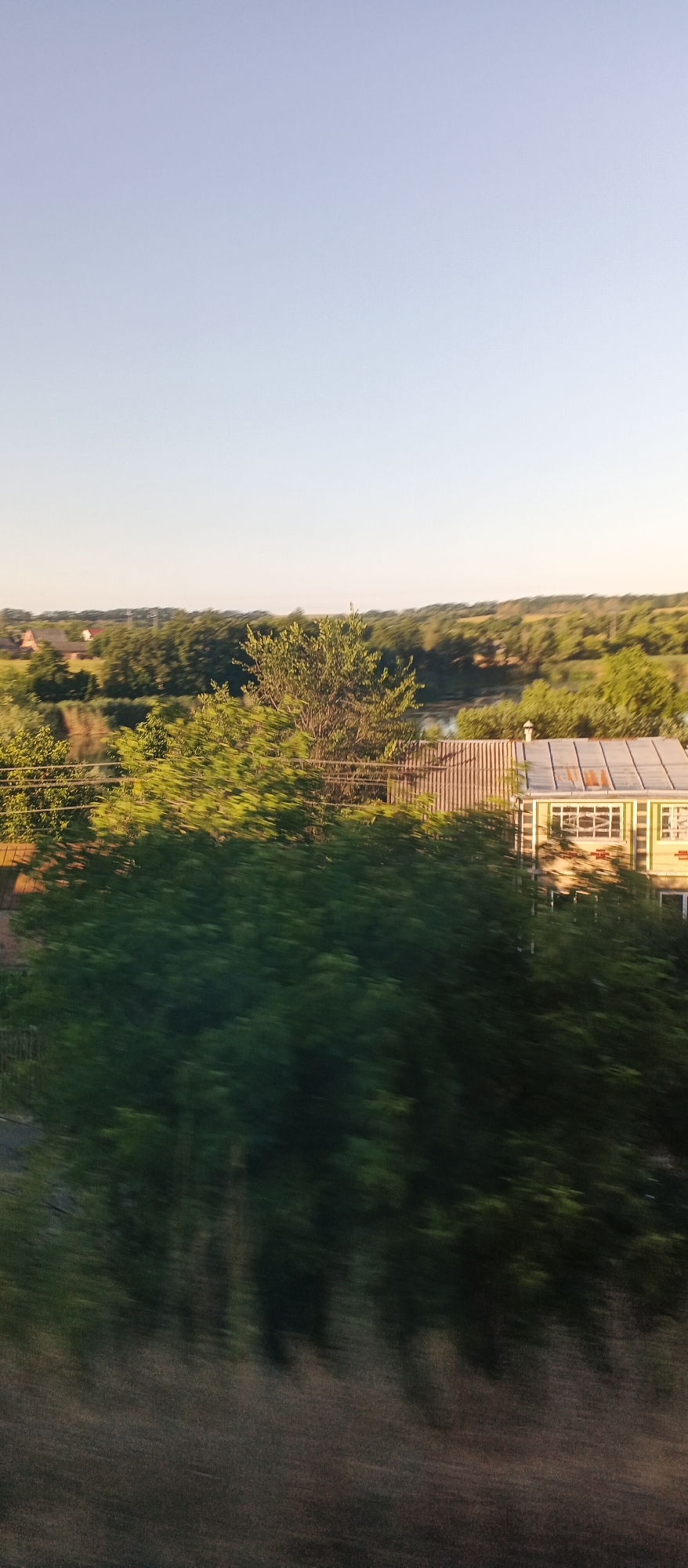 The image depicts a rural landscape at dusk, possibly in or near Khlystunivka, Ukraine. The sky is a clear, pale blue, transitioning to a lighter hue near the horizon. In the middle ground, a cluster of trees forms a dense line, with a single, larger tree standing out in the center. To the right of the trees, a small, light-colored building with a corrugated metal roof is visible. The building appears to have several windows with decorative grilles. In the foreground, a blurred swath of dark green foliage suggests a hedge or dense undergrowth, indicating movement or a shallow depth of field. The background is filled with rolling hills covered in trees, with a few scattered houses visible in the distance. The overall impression is one of a tranquil, pastoral setting.