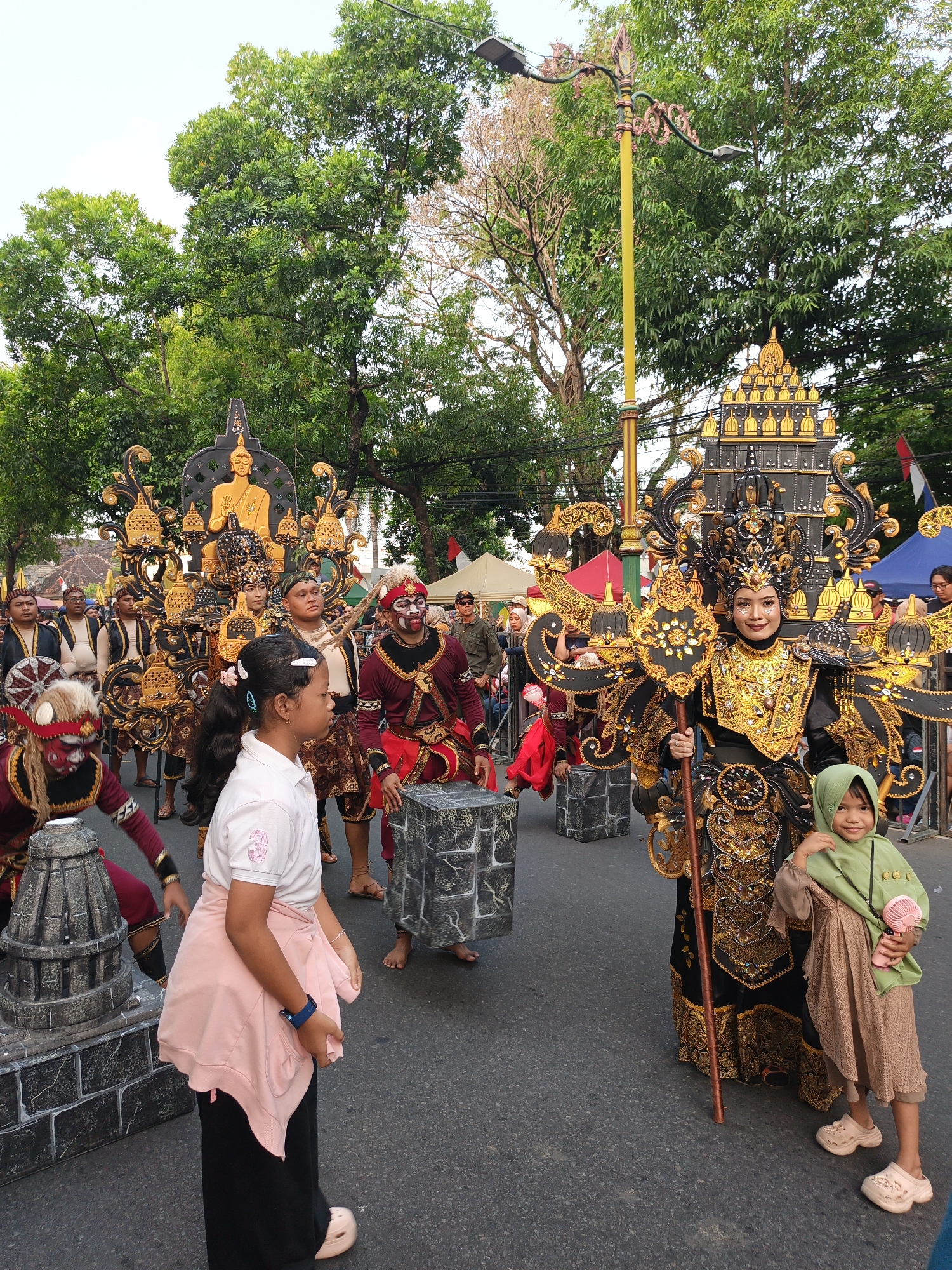 The image depicts a street parade in Klaten, Indonesia, likely during a cultural festival. The central focus is a group of performers in elaborate, traditional costumes. One prominent figure is adorned in a black and gold headdress and ornate outfit, holding a staff. To her left, a young girl in a pink shirt and black pants stands facing the side. To the right of the main performer, another young girl in a light green headscarf and brown dress holds a pink fan.
Several other performers are visible, some wearing red and maroon outfits with masks or face paint. They are positioned around large, decorated floats or structures that appear to be part of the procession. In the background, trees line the street, and a lamppost with ornate details is present. Tents or marquees can be seen in the distance, suggesting a festive atmosphere. The ground is paved asphalt, indicative of a city street. The overall scene suggests a lively and colorful cultural event.