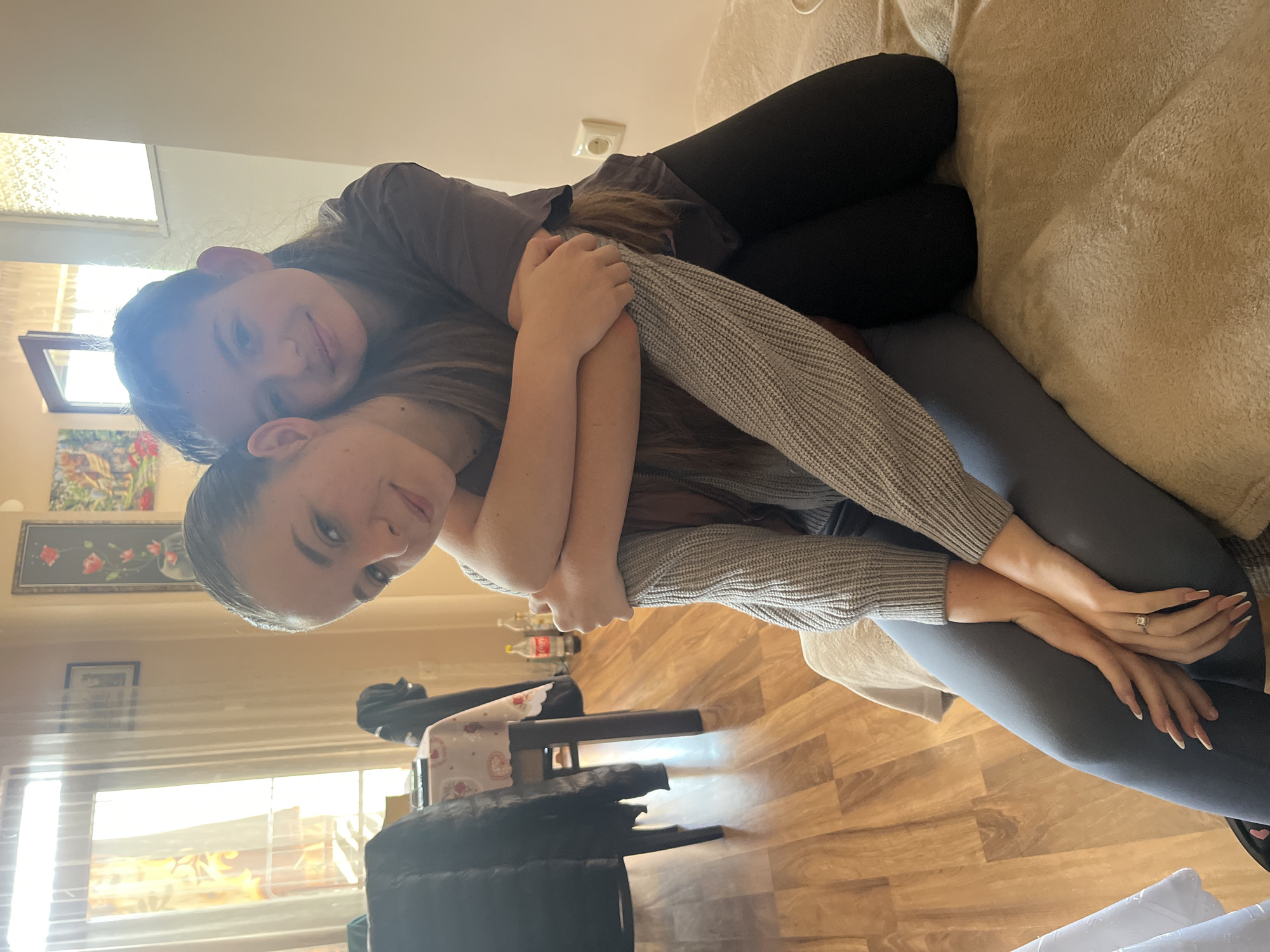 Two young women are embracing on a light-colored couch. One woman is wearing a grey-brown top and black leggings and is lying on the couch with the other woman behind her. The second woman wears a grey sweater and grey leggings. They are indoors, as indicated by the window, curtains, wall hangings, and hardwood floor. There is a chair and table visible in the background, along with water bottles. Based on the location in the city of Nova Zagora, Bulgaria, the image was likely taken in a residential setting.