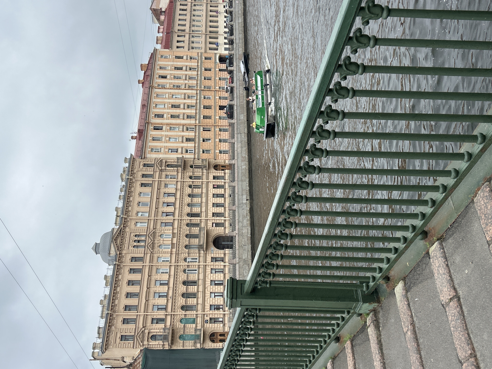 The image captures a view of a canal with buildings and a bridge railing in the foreground. The buildings lining the canal are of a classic architectural style, with multiple stories, numerous windows, and ornate facades in shades of beige and orange. Some buildings have red-tiled roofs. The canal water is a dark, murky brown, with ripples indicating movement.
In the canal, a green and white tour boat is visible, carrying several passengers. The boat has a covered section with windows and an open rear deck. Behind the tour boat, another sleek, dark boat is moving in the same direction. Along the opposite bank of the canal, cars are parked and driving on a street that runs parallel to the water.
The foreground is dominated by a dark green metal bridge railing. The railing features a decorative pattern with vertical bars and ornate elements at the top. To the left of the railing, the steps of the bridge are visible, constructed with grey concrete and bordered by rough-textured stone. The sky is overcast, contributing to the subdued lighting of the scene. The overall impression is of an urban waterway scene, likely in a European city.