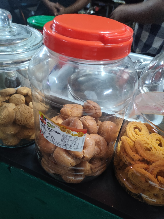 Three clear plastic jars filled with various snacks are displayed on a dark surface, likely a counter in an indoor setting. In the foreground and center, a large clear jar with a bright red lid holds numerous light brown, irregularly shaped, glazed snacks. A label on this jar identifies the product as "RADHAS SEMIYA CHIKKI," weighing 250 GM and containing 20 pieces. The manufacturing date is 20-09-2023, and the expiry date is 19-12-2023. The label also mentions "RADHAS FOOD PRODUCTS" located in Perumon P.O., Kollam Dt. 691 601, Kerala, India.
To the left of the central jar, another clear glass jar with a transparent lid is filled with smaller, round, light brown cookies or biscuits. To the right, a portion of a third clear plastic jar is visible, containing bright yellow, spiral-shaped savory snacks, commonly known as Murukku or Chakli in India.
In the blurred background, parts of two individuals are discernible. One person's arm and torso, clad in a plaid shirt, are visible behind the red-lidded jar. Further back, another pair of hands can be faintly seen. The scene suggests a retail environment such as a local shop or a market stall in Munderi, India, where packaged food items are sold. The lighting appears to be artificial and diffused.