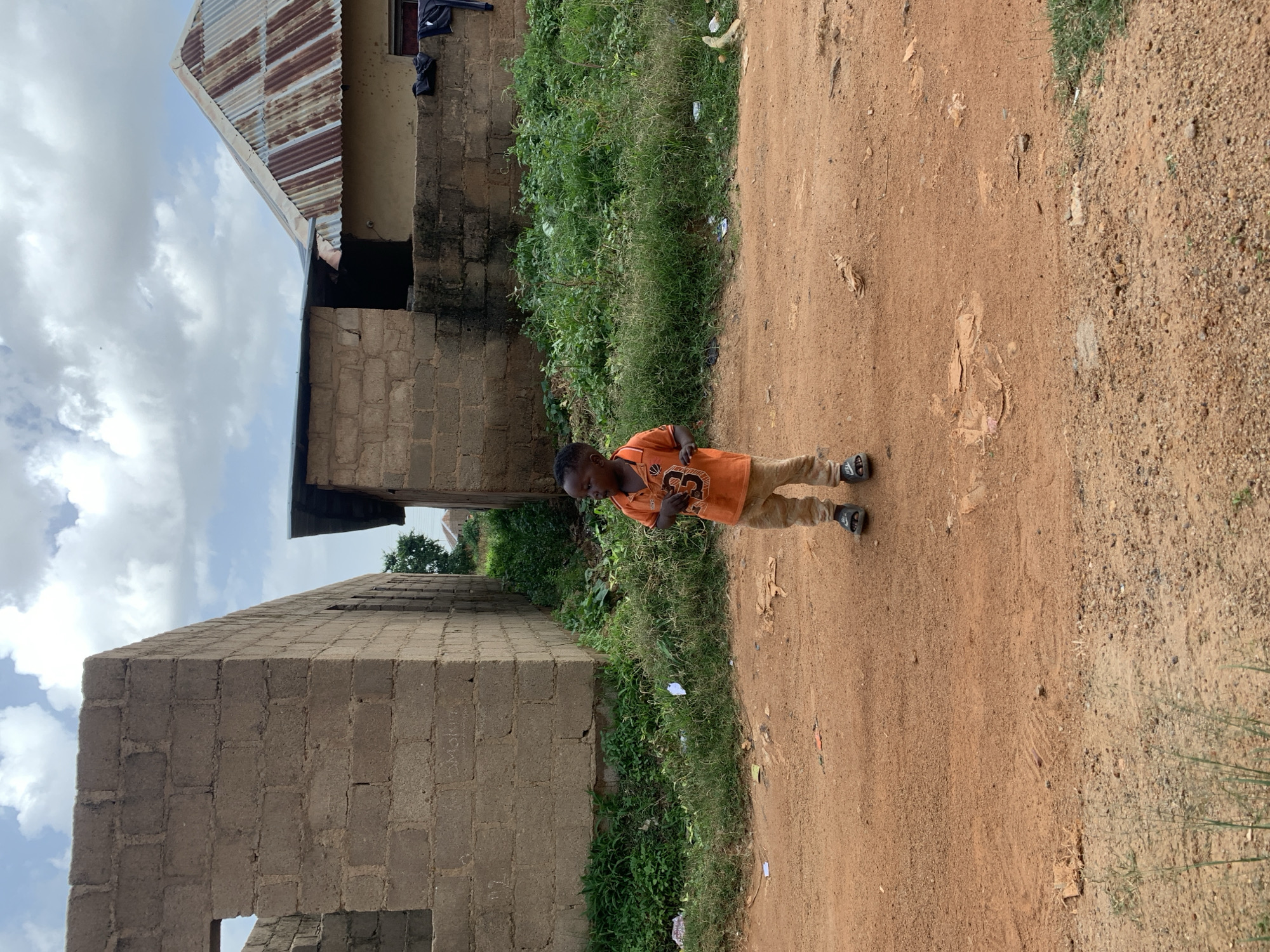 A young boy stands on a dirt path in front of a brick building. The boy wears an orange t-shirt with a graphic on it and light brown pants. He is facing left and looking down at his hands. He has dark hair and dark skin. Behind him, a building is under construction with rough brick walls and a corrugated metal roof. To the right of the building, there is a patch of green grass. The sky is cloudy. The scene appears to be in Du, Nigeria. The time of day is likely daytime, given the lighting. There is no discernible text in the image.