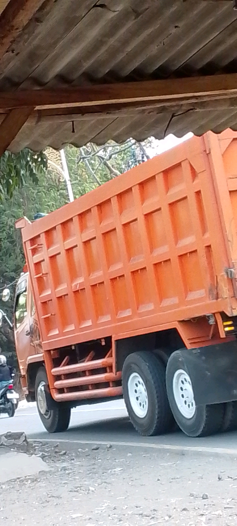 In Cilaku, Indonesia, an orange dump truck is parked under a wooden overhead structure. The truck's bed is filled with visible compartments, and it has dual rear wheels with white rims. To the left of the truck, a motorcycle with a rider wearing a white helmet is visible. The environment appears to be a roadside, with trees and foliage in the background, and a paved road surface with some debris.