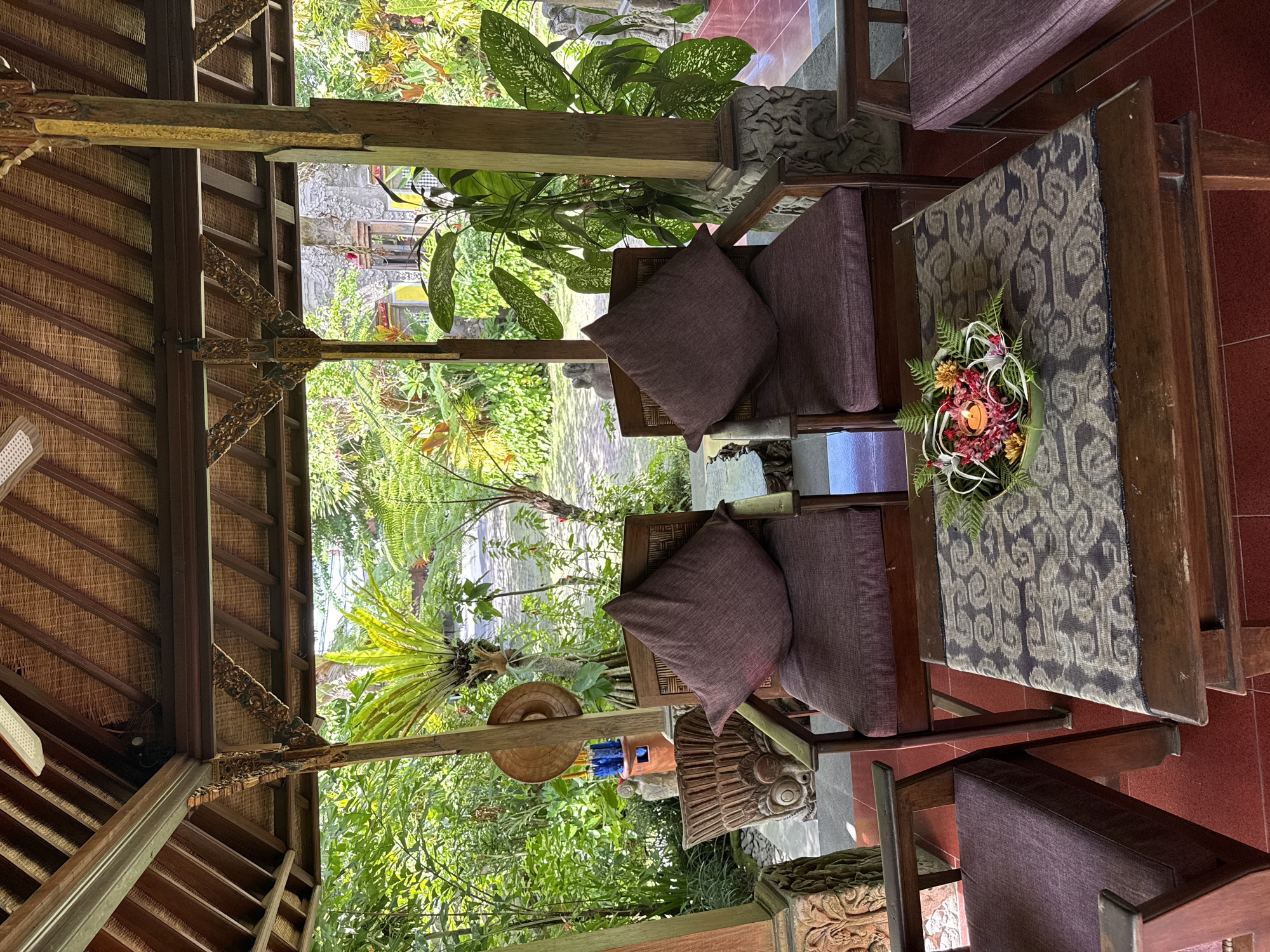 The image depicts an outdoor seating area in Ubud District, Indonesia, featuring two wooden chairs with purple cushions and decorative pillows, flanking a wooden coffee table. The table is adorned with a centerpiece of fresh flowers and a lit candle. In the background, lush green foliage and tropical plants create a serene atmosphere. The architecture suggests a traditional Balinese style, with a thatched roof and intricately carved wooden elements. The ground is paved with red tiles. The scene is well-lit by natural sunlight, casting soft shadows.