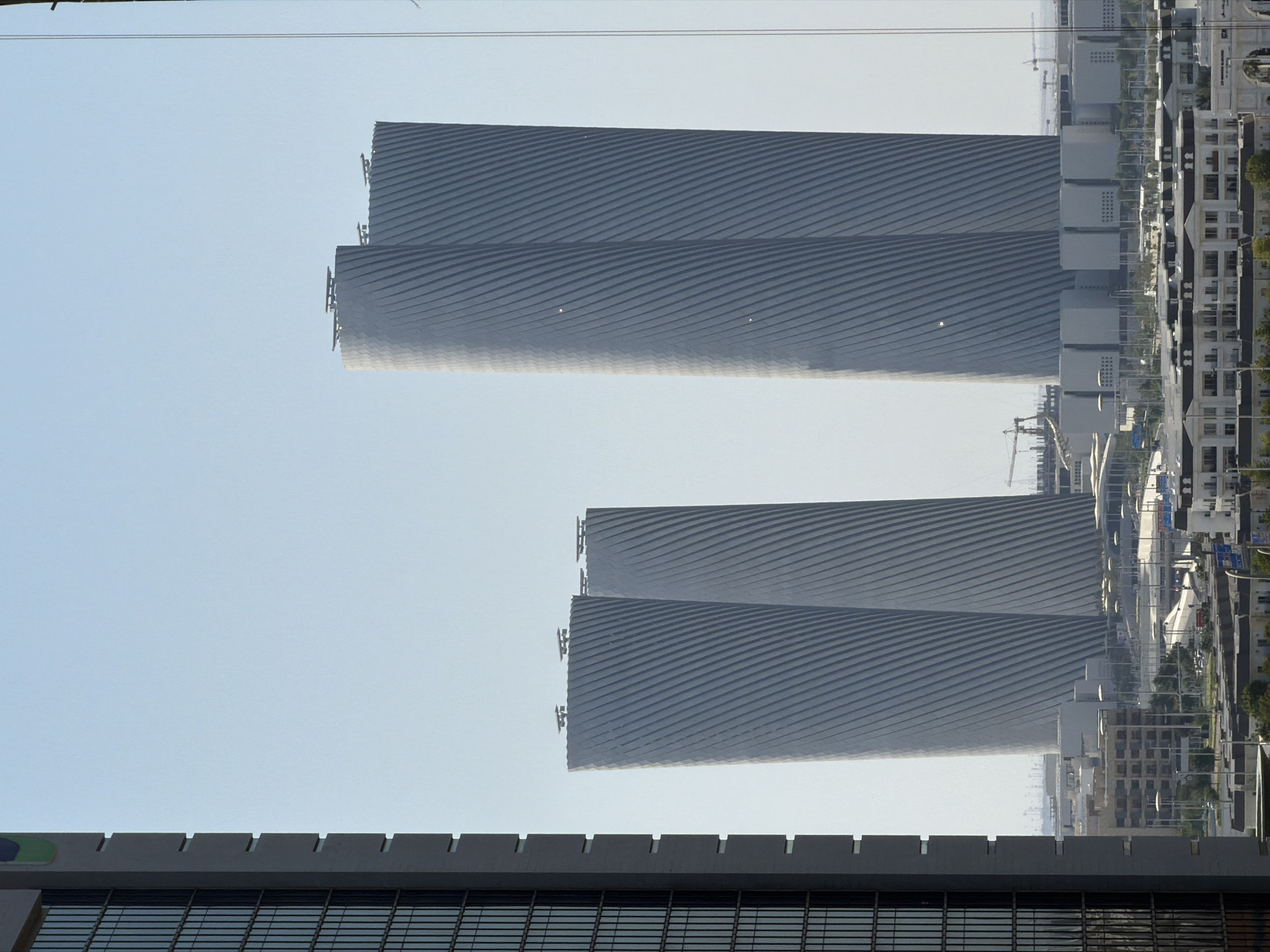 An exterior, eye-level, daytime, medium shot captures a cluster of modern skyscrapers under a clear, pale blue sky. The foreground is dominated by the left side of a dark gray, rectangular building with a grid of small, dark windows. To the right of this building are two pairs of tall, cylindrical skyscrapers with a distinctive ribbed texture that twists diagonally up their surfaces. The two closest towers are identical in design, appearing as a single, wider structure from this angle. The tallest skyscraper is to the far right. Each tower has a flat top with several rectangular structures or platforms. In the middle ground, a lower-rise cityscape unfolds, featuring buildings with flat roofs and multiple rows of windows, interspersed with streetlights and vegetation. Several construction cranes are visible in the background, suggesting ongoing development. The overall scene depicts a contemporary urban environment.
