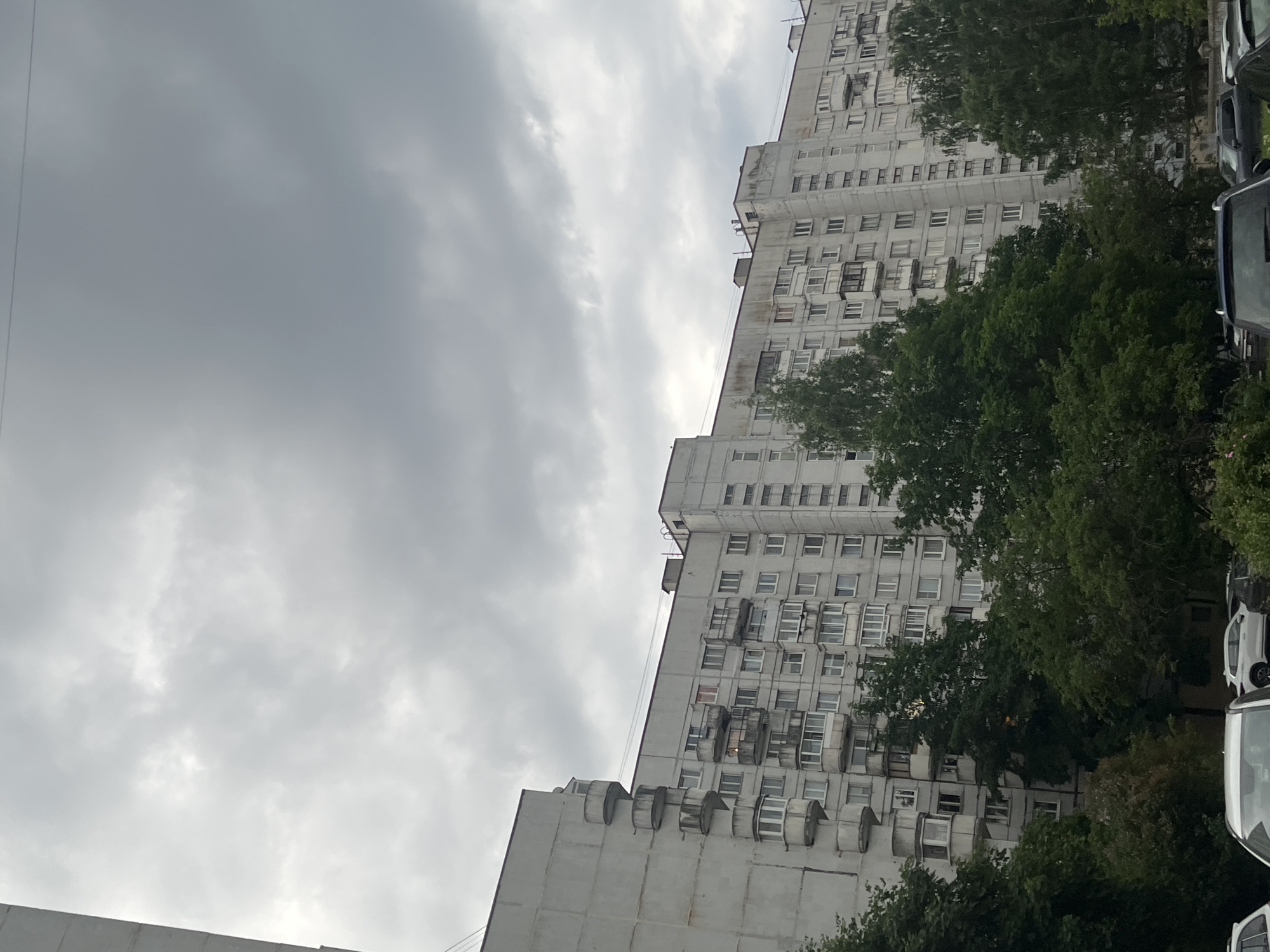 The image depicts a large, multi-story residential scene in Saint Petersburg, Russia, under an overcast sky. The primary subject is a lengthy, light-grey, concrete apartment building of a type common in Soviet-era mass housing construction. The building features numerous rectangular windows, some with enclosed, glazed balconies, and others with open balconies. A distinctive architectural detail is visible on the leftmost section of the building, where several semi-circular, open balconies are stacked vertically. The building's facade shows signs of weathering and discoloration, particularly beneath the roofline and around some windows. A reddish curtain or blind is visible in one window on the middle-left section, and a warm interior light illuminates another window slightly to its right.
The sky is uniformly grey and heavily clouded, indicating an overcast day. Dense, mature deciduous trees with lush green foliage occupy the middle and lower foreground, partially obscuring the lower floors of the apartment building and the ground level. Below the trees, parts of several parked cars are visible, including the front hood of a white SUV on the left and various dark-colored vehicles on the right. No people are visible, and no specific actions or interactions are depicted.
The architecture, particularly the panel construction and the design of the balconies, is characteristic of the **137-series apartment blocks** widely constructed in Saint Petersburg (then Leningrad) during the Soviet era, predominantly from the mid-1970s onwards. This type of residential block is common in the city's large residential "sleeping districts," such as **Kupchino, Ozerki, Grazhdanka, and areas around Prospekt Prosvescheniya**. While a precise street address cannot be determined from the visual information alone, the scene is unmistakably representative of these established residential neighborhoods within Saint Petersburg.