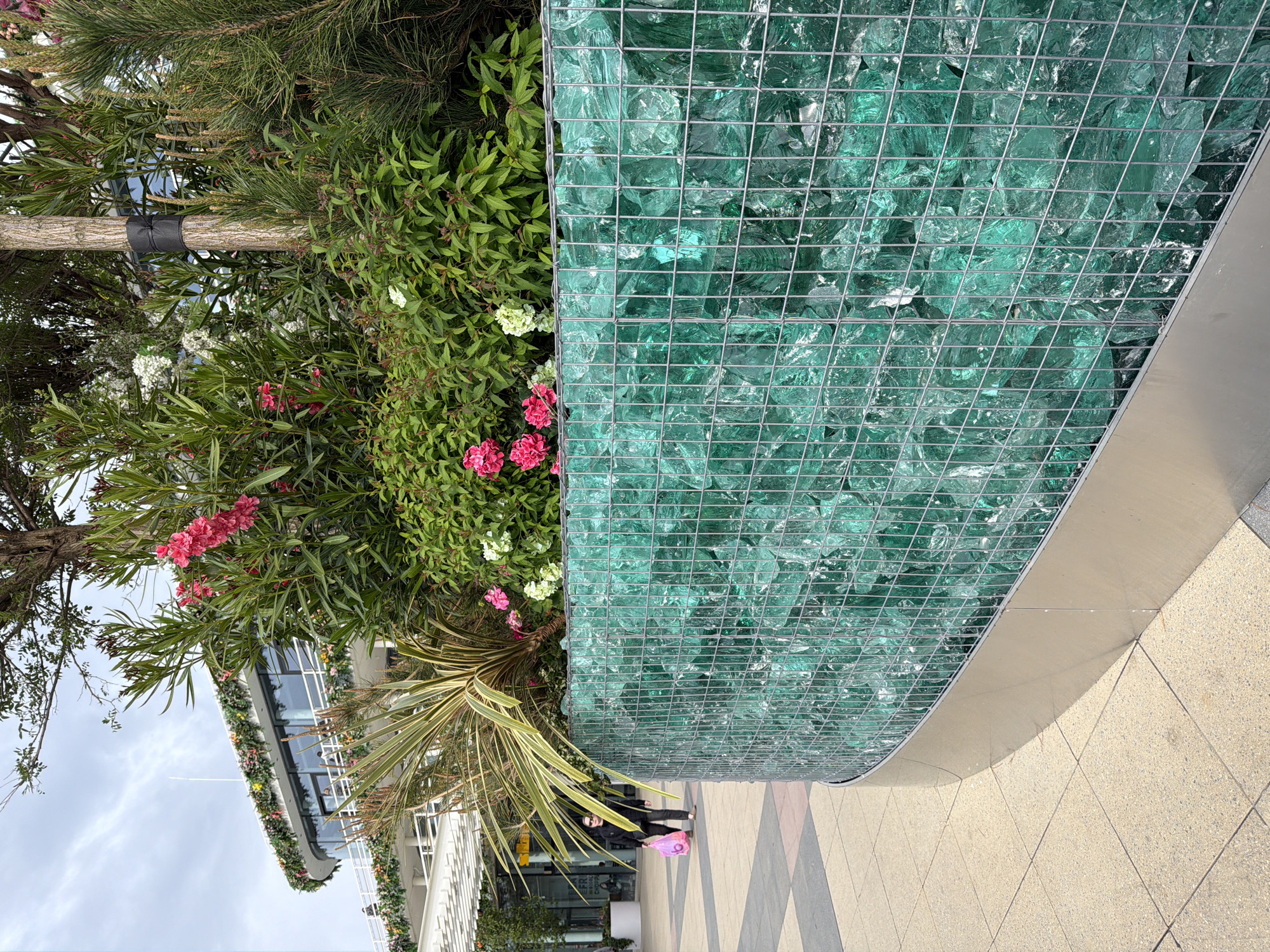 The image depicts an exterior scene, likely a public space or commercial area. On the right, a structure made of large, light green glass pieces held together by a metal wire mesh is visible. Adjacent to this, a variety of plants, including leafy bushes with pink and white flowers, and a palm-like tree, create a green backdrop. In the background, a multi-story building with glass windows is visible, and a cloudy sky suggests an outdoor setting. A person is standing in front of the plants. A pink bag is visible near the person. The location is not specifically identifiable as Moscow, Russia from the visual information provided.