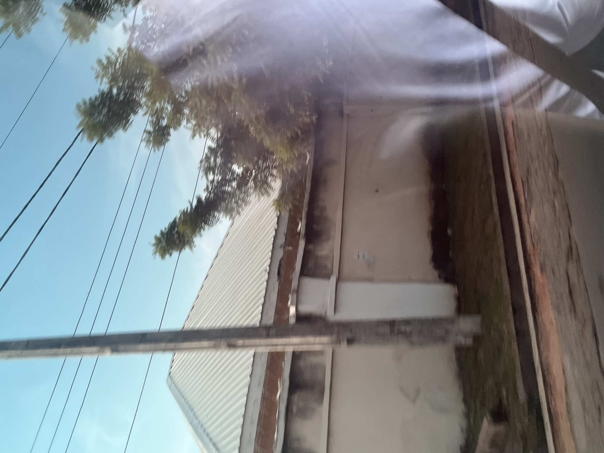 The image displays a view of a building with a corrugated metal roof, possibly a workshop or storage area, in Jalingo, Nigeria. The building has a light-colored exterior wall, and what appears to be a gray metal pipe or conduit runs horizontally across the front. In the background, trees with green foliage are visible against a bright blue sky. Several dark power lines stretch across the frame, suggesting an overhead electricity supply. The image is taken at an angle, possibly from a vehicle or another elevated perspective, with reflections visible on what might be a window or a screen, obscuring some details and adding a hazy quality to the scene. The lighting suggests it is daytime, likely sunny. The overall impression is of an outdoor industrial or semi-industrial setting. There is no visible text in the image.