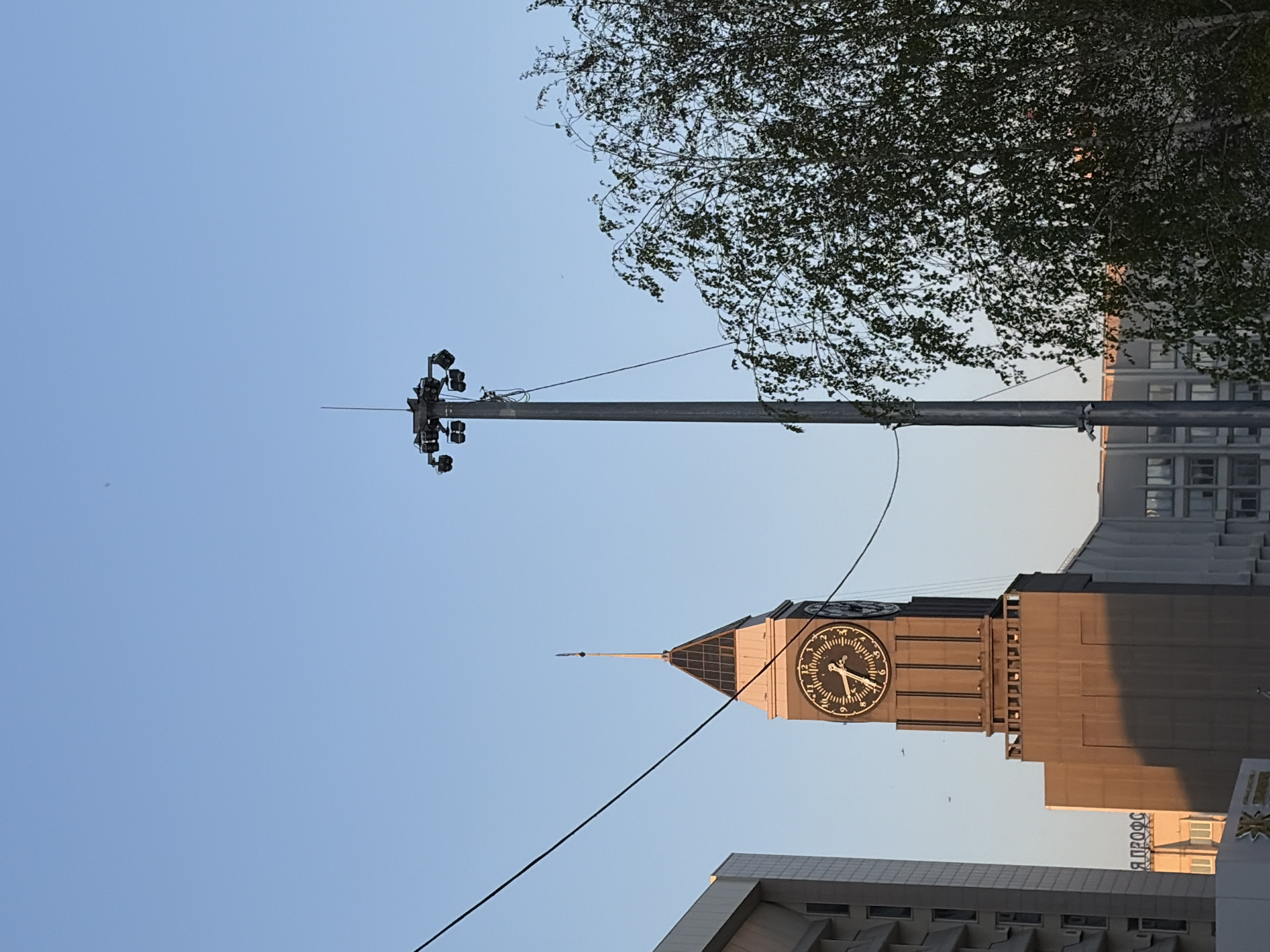 The image shows a clock tower in Krasnoyarsk, Russia, viewed from below, with the tower dominating the right side of the frame. The clock face displays a time just before 8:00 AM. A tall, light-studded pole extends vertically on the left, partially obscured by the tree branches in the top right. The sky is visible in the background. Buildings surround the clock tower. A wire runs diagonally across the foreground.