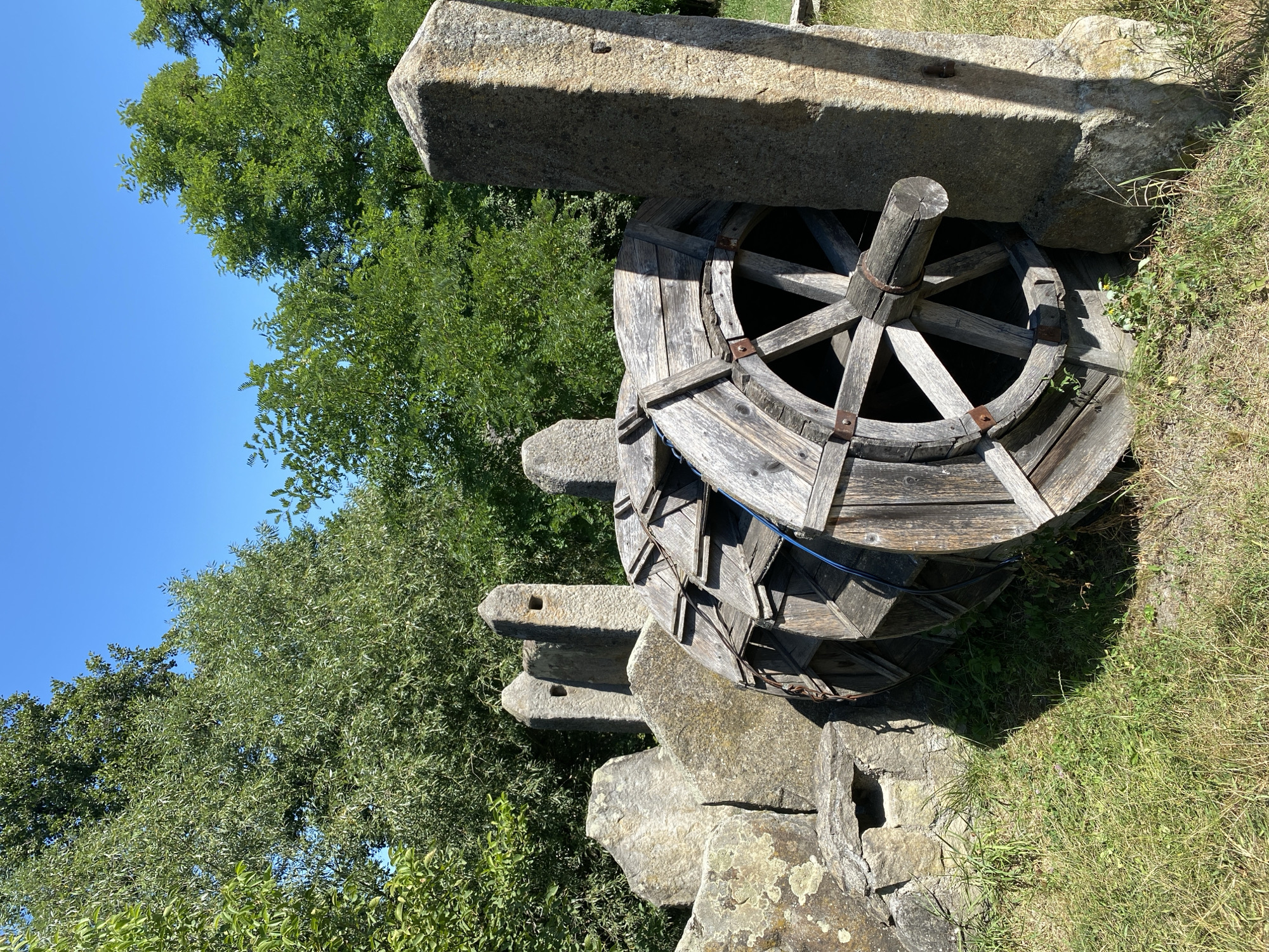 The image shows a large, wooden water wheel resting on the grass, partially supported by a stone structure. The water wheel is constructed from weathered planks and has a visible axle. To the left of the water wheel, there are several large, rough-hewn stones, some of which have openings or indentations. Behind these stones, two pointed stone structures, resembling small obelisks, rise upwards, each with a square opening. To the right of the water wheel, a tall, rectangular stone pillar stands upright. The background is filled with lush green trees and a clear blue sky. The overall scene appears to be outdoors in a grassy area, possibly a historical site or park, given the rustic nature of the objects and the presence of stone structures. The location is identified as Busha, Ukraine. There are no people visible in the image.