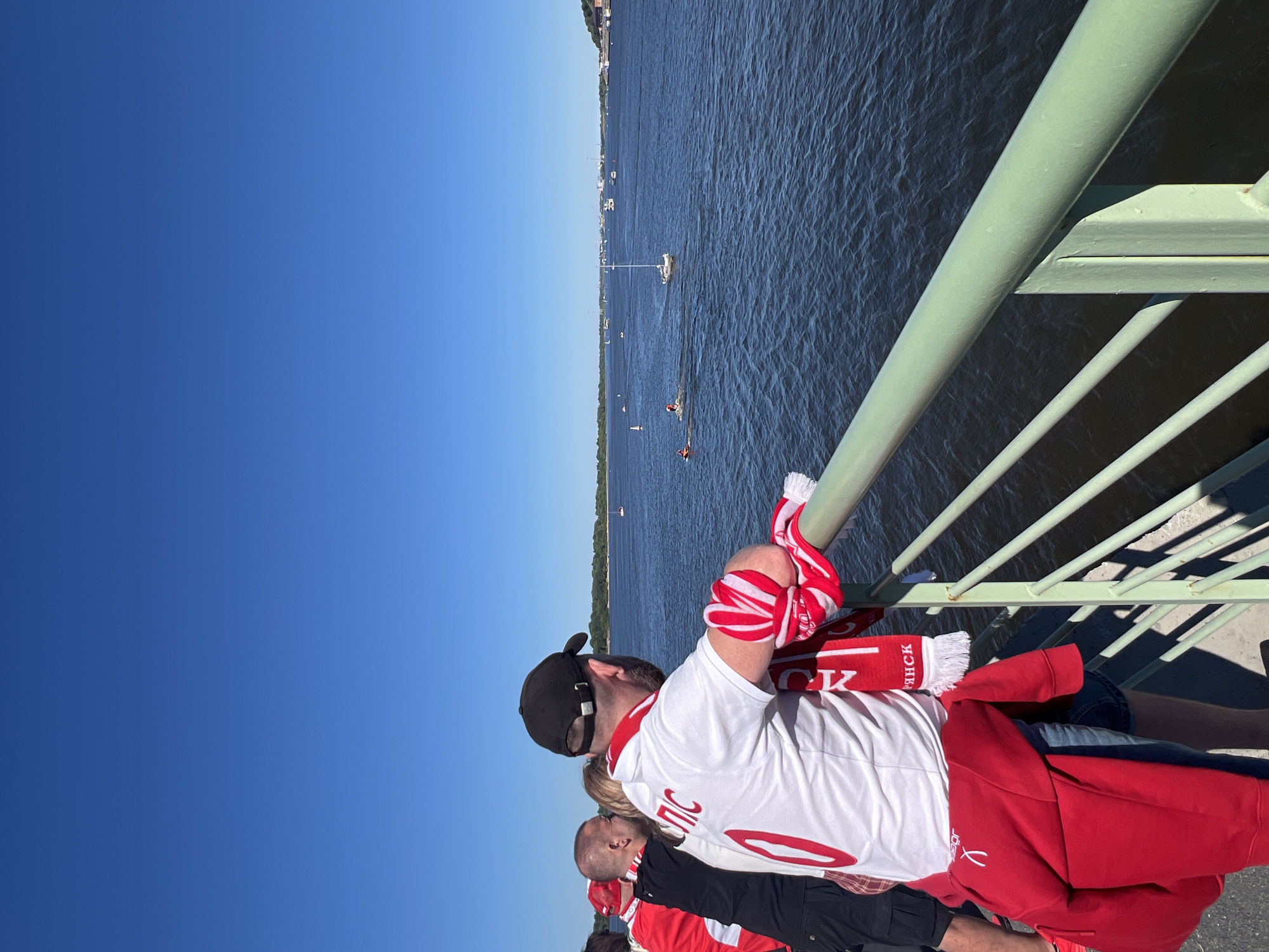 The image shows a group of people wearing red and white scarves and shirts, likely sports fans, standing on a bridge over the Volga River in Nizhny Novgorod, Russia. The sky is clear and blue, and the water is dark with visible ripples. Several small boats and a sailboat are visible on the river. In the foreground, a man in a white shirt with red lettering and a red scarf is leaning on a green railing. Another man in a black shirt and a person in a red shirt are also visible in the background. The overall scene suggests a bright, sunny day and a sporting event or a leisure activity taking place on the water.
