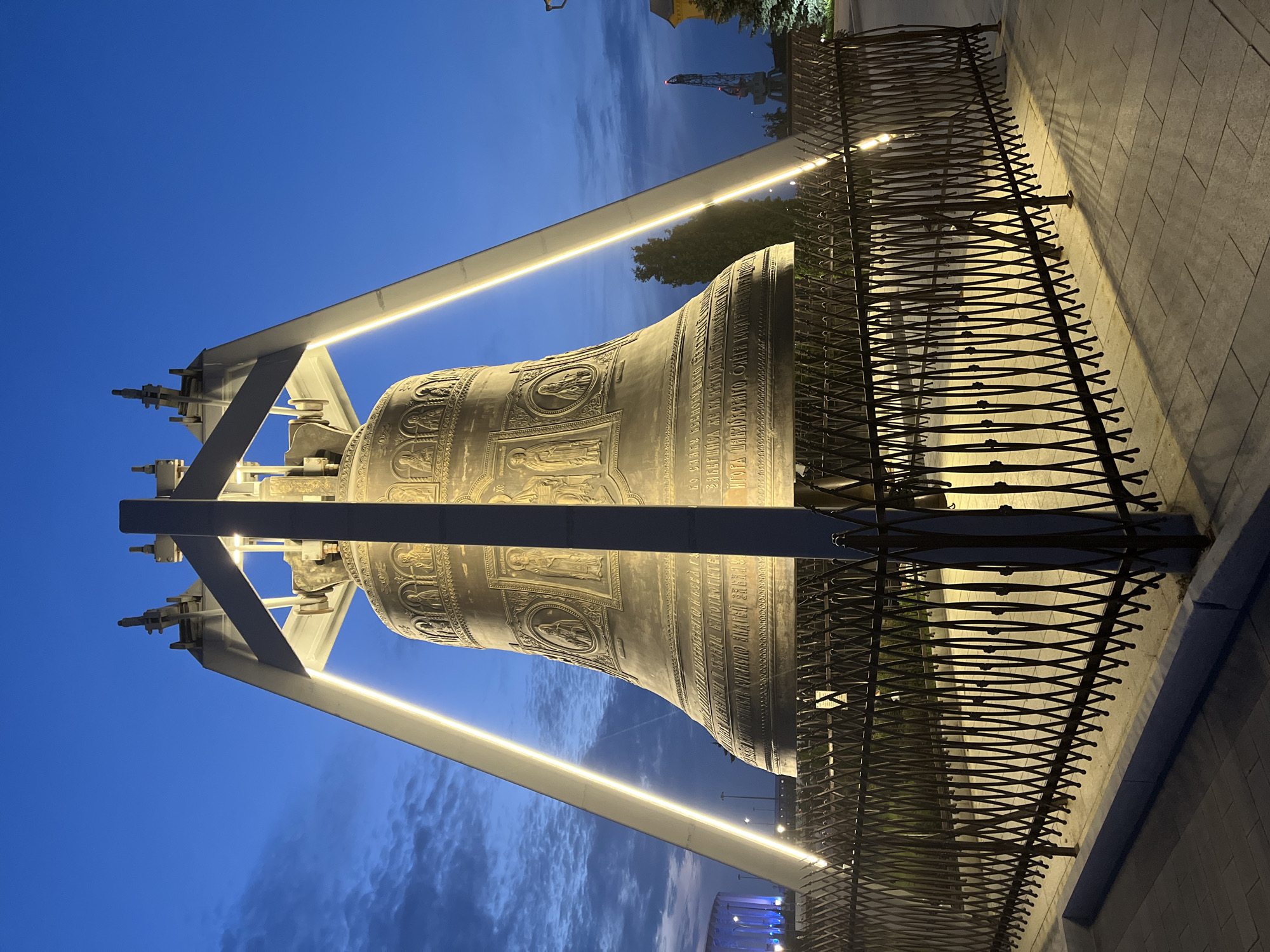 The image shows a large bell, likely a bell tower, in Nizhny Novgorod, Russia. The bell is made of bronze and is ornately decorated with bas-relief sculptures of saints and religious figures, as well as inscriptions. It is supported by a large metal frame, which is illuminated by vertical LED lights. The bell is enclosed by a decorative black metal fence. The scene is set at dusk, with a clear blue sky with some clouds. In the background, there are some trees and buildings, including a structure that appears to be a stadium or arena with blue lighting. A tall industrial crane is visible in the distance. The ground is paved with interlocking concrete blocks. There are no people visible in the image.