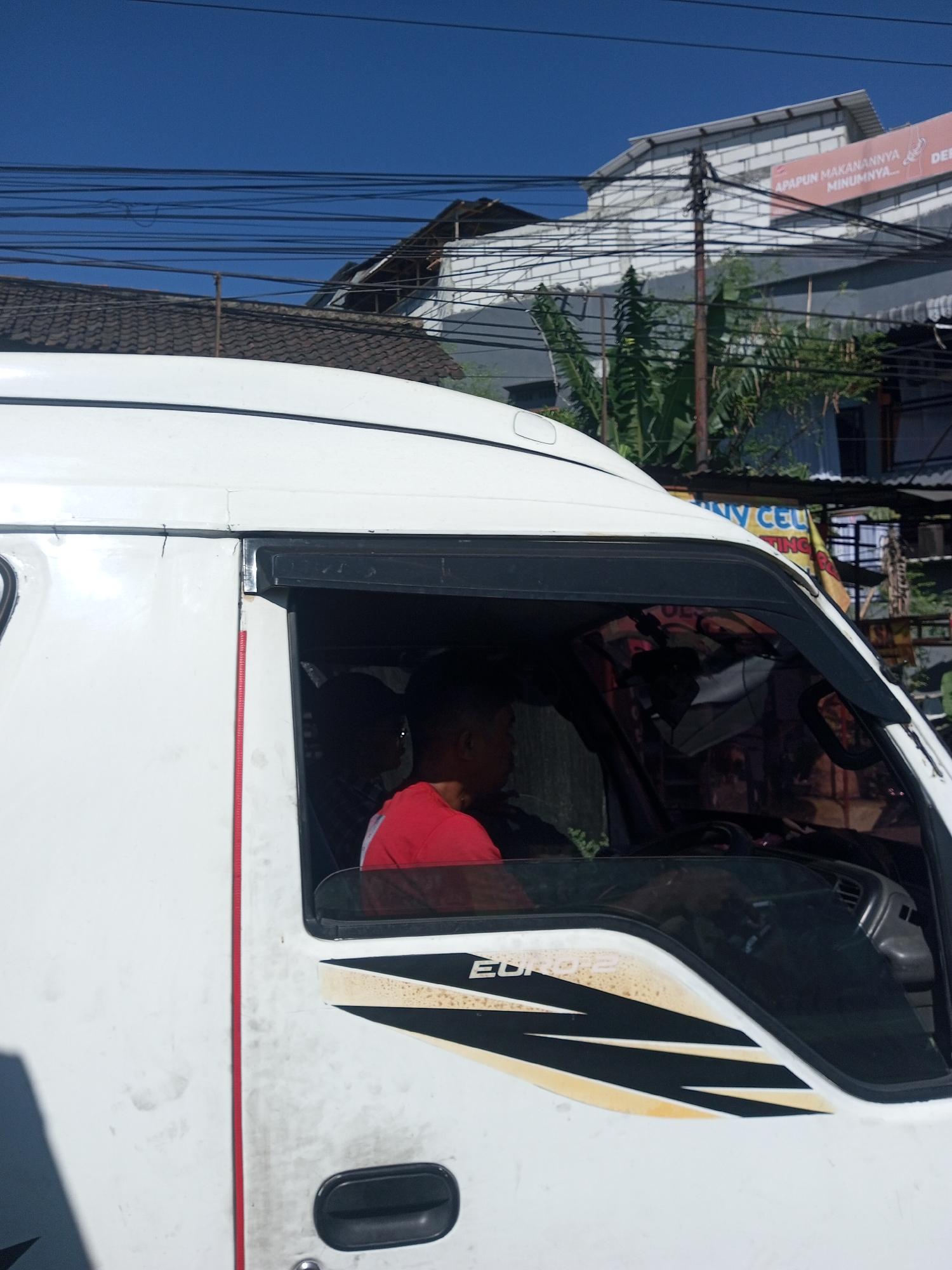 A white van with a black stripe decal reading "EURO2" is parked on a street. A man wearing a red shirt is visible inside the van, in the driver's seat. The street appears to be in a city, with buildings and power lines in the background. The sky is clear and blue. The scene suggests an ordinary day in a built-up area.