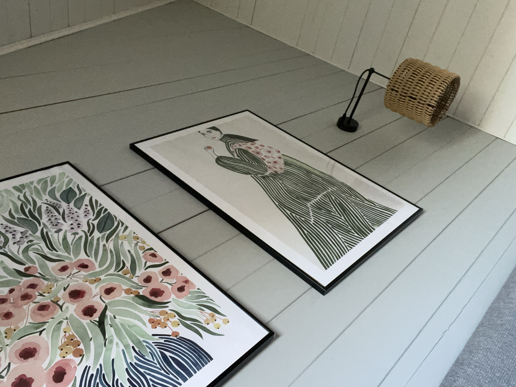 The image shows two framed artworks leaning against a wall with light blue horizontal paneling. A small, woven lamp with a black stand sits to the right of the rightmost artwork. The left artwork features a watercolor painting of flowers in shades of pink, green, yellow, and blue. The right artwork features an illustration of a person in a green striped dress holding a red object. The environment appears to be an interior setting, likely a room in a home or apartment. The city of Литвиново, Russia, cannot be definitively determined as the location from the visual content of the image.