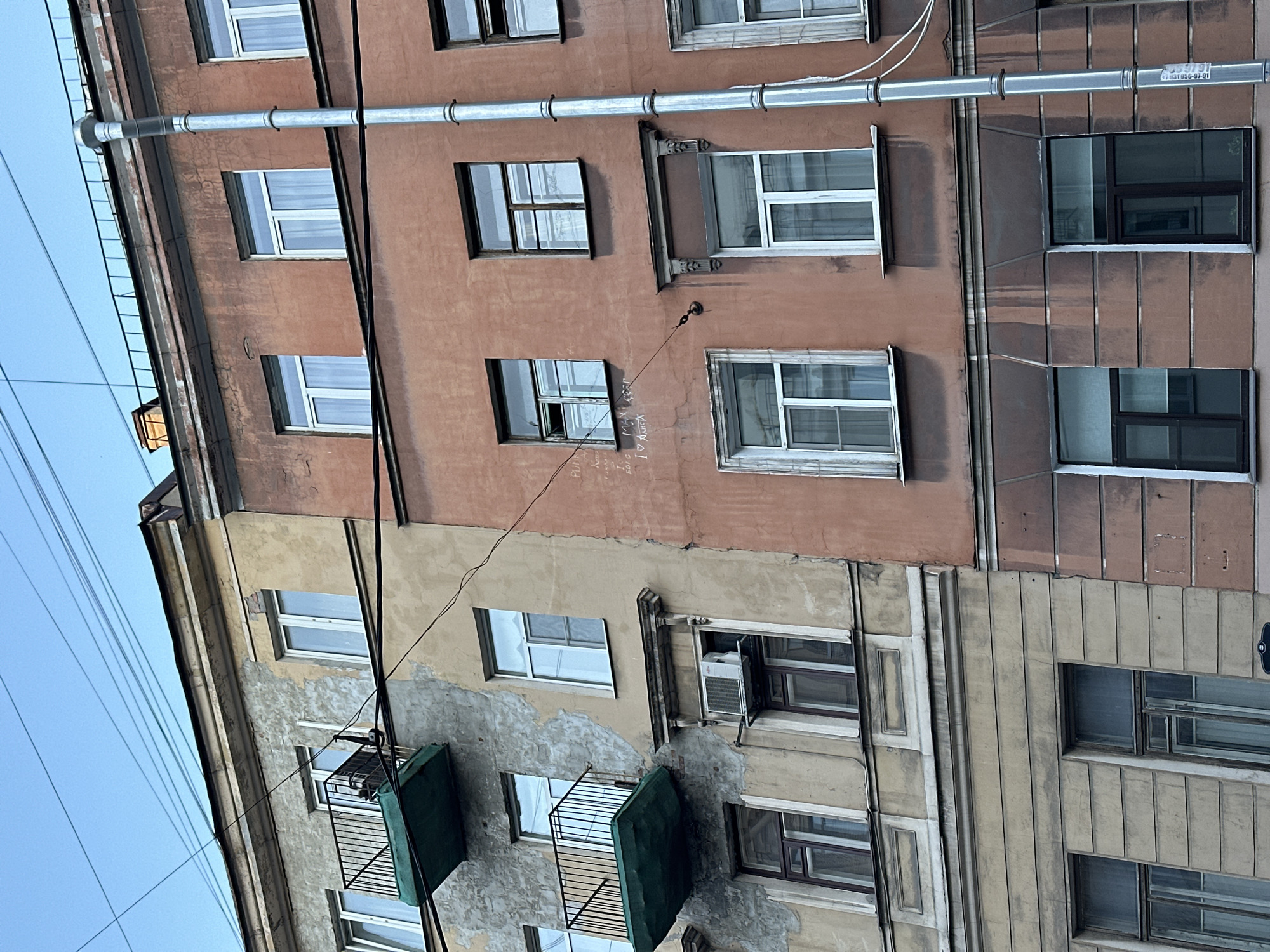 The image depicts a section of a building facade in Saint Petersburg, Russia. The building consists of at least two distinct sections, differentiated by color and architectural style. The upper section is painted in a reddish-brown hue, with simple rectangular windows. A drainpipe runs horizontally along the facade. The lower section is a lighter, beige color and features more ornate detailing around the windows, including fluted columns. Some windows have air conditioning units installed and small, green-covered balconies. Electrical wires are visible against the sky. There are no people visible in the image.