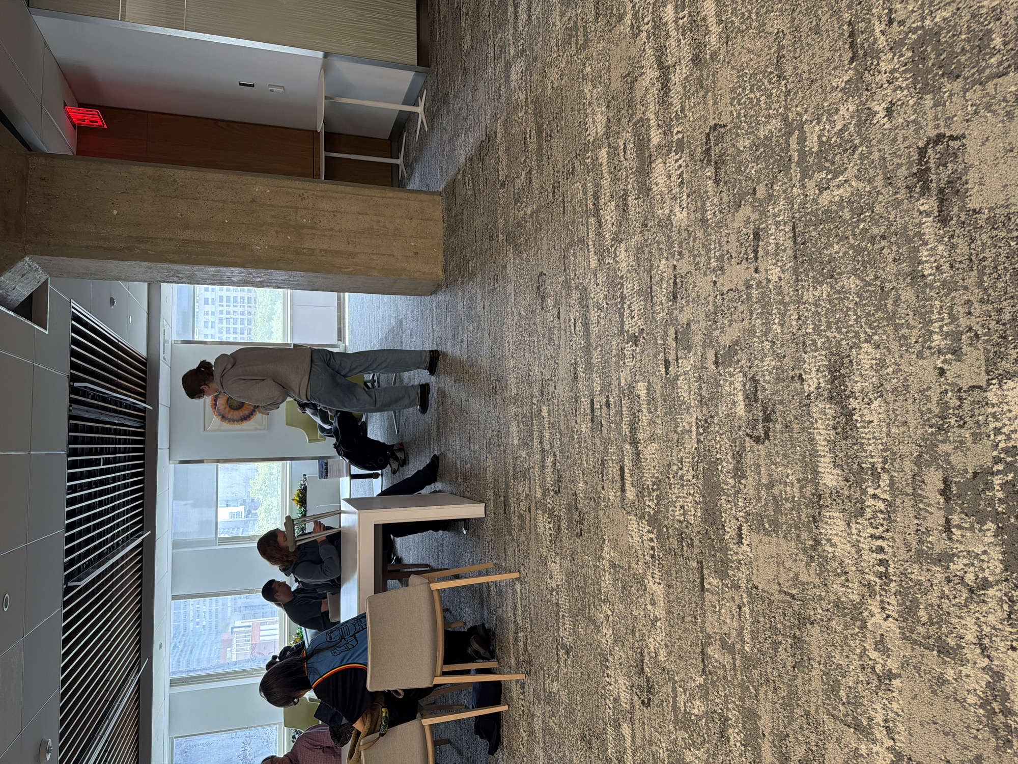 The image depicts an interior setting, likely a study area or library in New York City, evident from the cityscape visible through the windows. A group of people are seated at a long table, engaged in various activities. Some appear to be studying or reading, while others are in conversation. A person wearing a gray hoodie is standing, leaning over the table, perhaps observing or engaging with the seated individuals. A pair of crutches lean against the table. The environment includes a textured carpet, large windows overlooking the city, and architectural elements such as a concrete beam and window blinds. There is an emergency exit sign visible in the upper left of the scene.