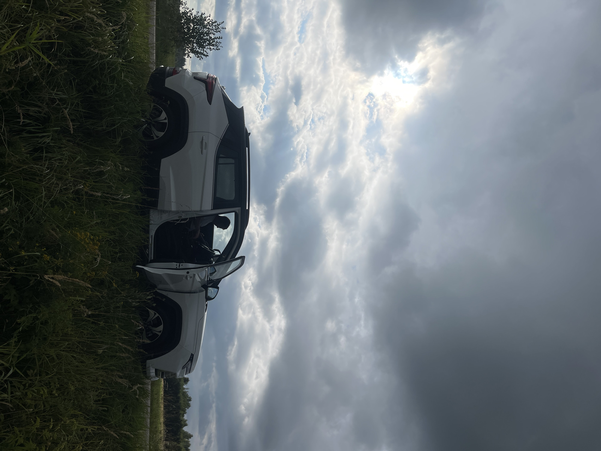 A white SUV is parked on the side of a road in a grassy field under a cloudy sky. The driver's side door is open, and a person is visible in the driver's seat, silhouetted against the interior light. The sky is dramatic with patches of sunlight breaking through dark clouds. A treeline is visible in the background. The image appears to have been taken during the day.