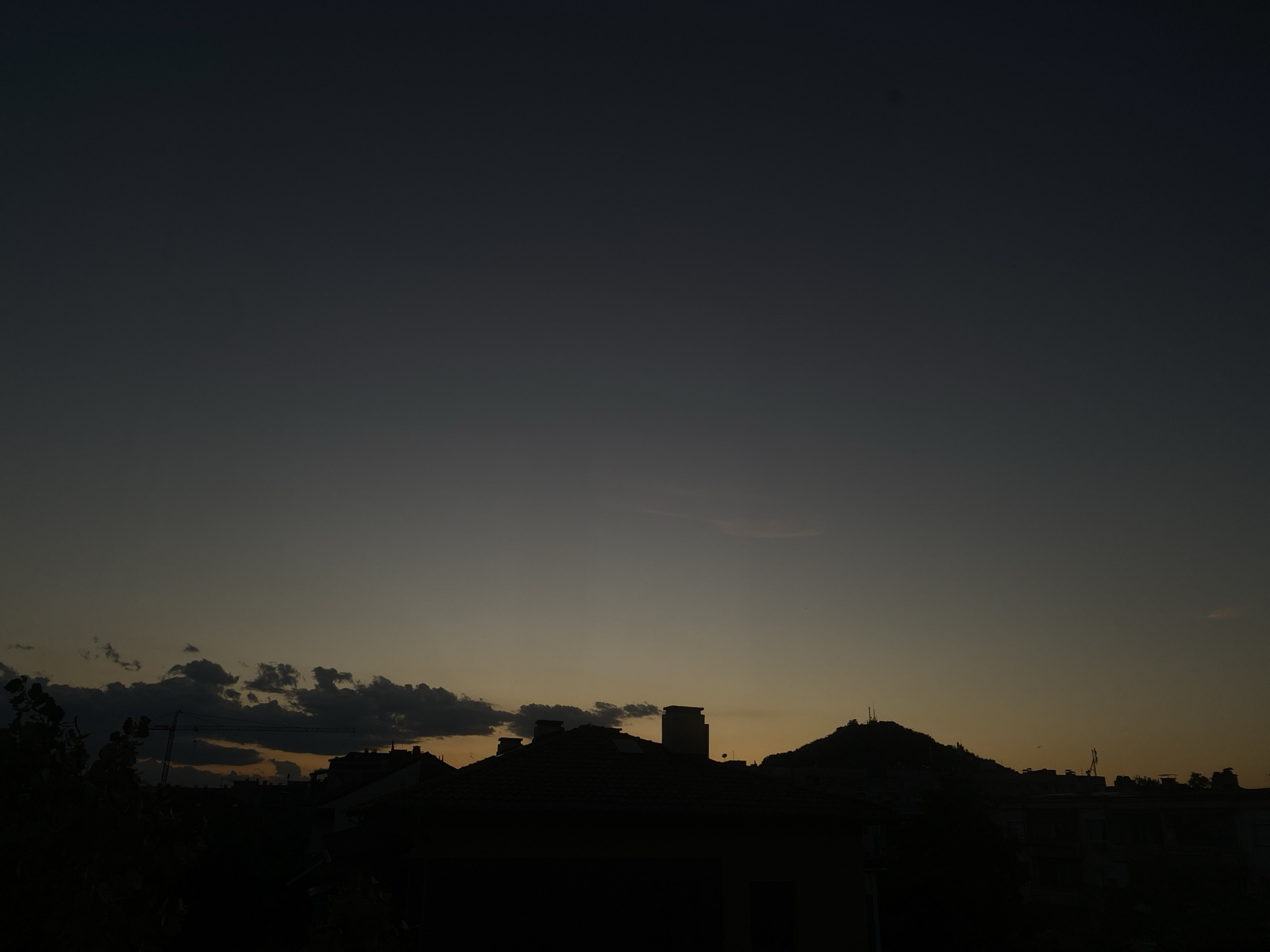 The image shows a cityscape at dusk, with the sky transitioning from deep blue to a pale orange near the horizon. Silhouetted against the fading light are buildings with distinct rooftops, chimneys, and a prominent hill topped with what appears to be a structure or antenna. Some clouds are visible, particularly towards the left side of the frame, with a few dark, dense formations and lighter wisps higher up. A construction crane is partially visible in the middle ground, adding to the urban landscape. The foreground is dominated by dark silhouettes of trees and more buildings, obscuring the ground level. The overall impression is one of a quiet evening in an urban environment.