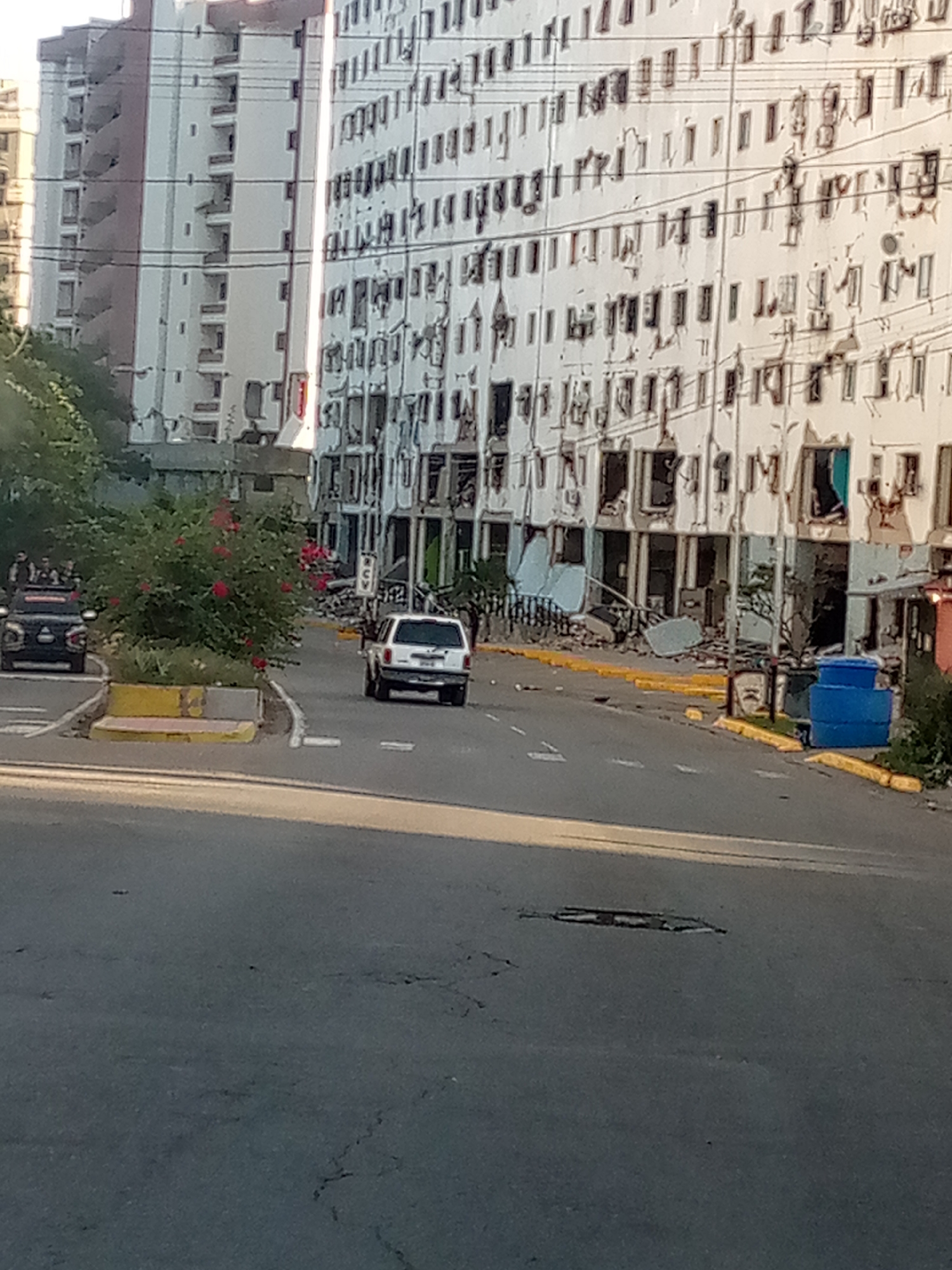 The image shows a street scene in Caraballeda, Venezuela, likely in the aftermath of a natural disaster. A large, white multi-story building on the right side of the street exhibits significant structural damage, with broken windows, crumbling facades, and debris scattered around its base. Yellow curbs line the street, and a blue plastic water tank is visible near the damaged building.
In the foreground, a dark-colored SUV is driving away from the viewer. Further down the street, a white SUV is also driving away. A dense green bush with red flowers is located on the left side of the road, partially obscuring a dark vehicle with several people in it. The road itself shows signs of wear and tear, with visible cracks and a pothole. The overall environment suggests a damaged urban area.