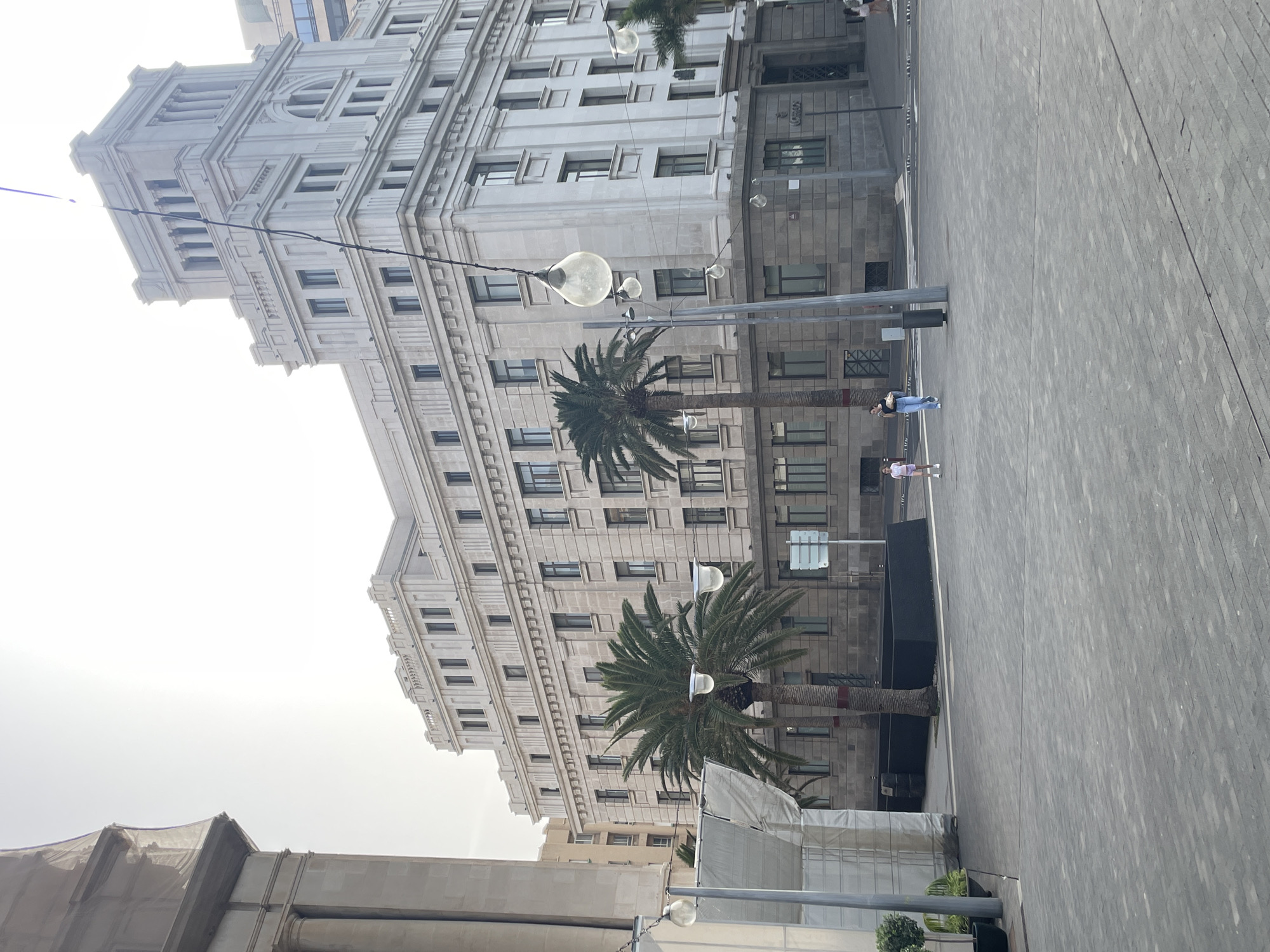 The image shows the Plaza de España in Santa Cruz de Tenerife, Spain, dominated by a large, multi-story building with ornate architectural details and numerous windows. Two palm trees stand in the plaza in front of the building. Hanging light fixtures are strung between poles. The ground is paved with gray stones or tiles. Two people are walking across the plaza. A building under construction is in the lower-left corner of the frame. The sky is overcast.