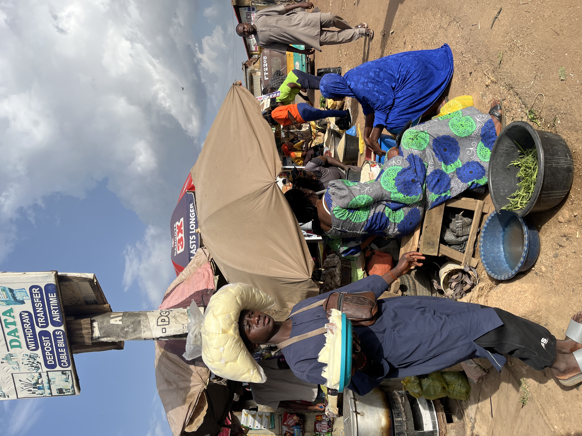 The image depicts an outdoor market scene in Rafin Par, Nigeria. In the foreground, a young individual wearing a navy blue robe and black pants carries a large bag of yellow produce on their head. They are also holding a light blue platter with slices of what appears to be cheese or butter. The person is wearing a brown satchel across their chest and light gray sandals.
Behind the person, various market stalls are visible, with people engaged in transactions and activities. Several individuals are seated and standing around stalls covered by umbrellas and tarps. One woman in a bright blue head covering and patterned dress is seated on the ground. Another woman in a green and blue patterned dress is also seated. A man in a grey tunic walks away from the viewer on the right side of the frame.
In the background, a signpost with various services listed such as "DATA", "WITHDRAW", "DEPOSIT", "TRANSFER", "CABLE BILLS", and "AIRTIME" is prominently displayed. A tricycle-like vehicle is parked behind some of the stalls. The ground is dusty, characteristic of an outdoor market environment. The sky is partly cloudy.