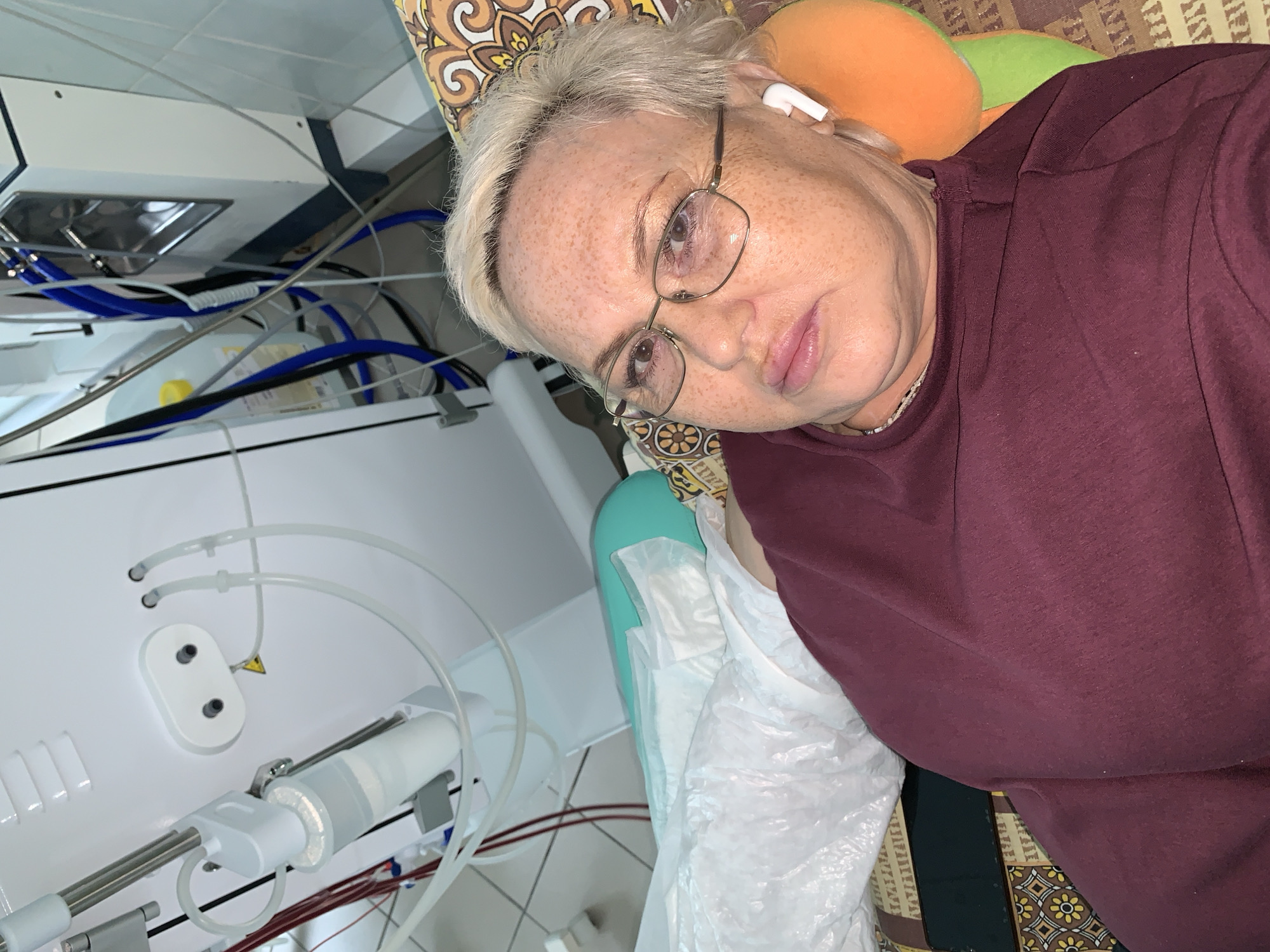A middle-aged or older woman with short, platinum blonde hair, prominent freckles, and rectangular glasses is depicted lying down, looking towards the camera. She wears a plain maroon long-sleeved top and a white wireless earbud in her right ear. Her head rests on a pillow with a distinct brown, yellow, and orange floral/geometric pattern, with an additional orange and green pillow visible behind it.
To her left, a complex white medical machine dominates the scene. This equipment features numerous clear, blue, and red tubes, and various white plastic components. Notably, there's a cylindrical clear component with what appears to be filters, and another white flat component with two holes and a small yellow triangular warning symbol. Visible red liquid within some of the tubes strongly suggests blood circulation, characteristic of a medical procedure like hemodialysis. The machine is connected to various wires and hoses, and the background behind it appears to be a light-colored, possibly tiled, wall. A blue-green medical pad and white plastic sheeting are also partially visible next to the woman.
The woman's expression is neutral, appearing calm or slightly pensive. The overall setting is clearly a medical environment, either a hospital room or a home adapted for significant medical care, located in Chaikovsky, Russia. The image is captured from a slightly overhead and tilted angle.