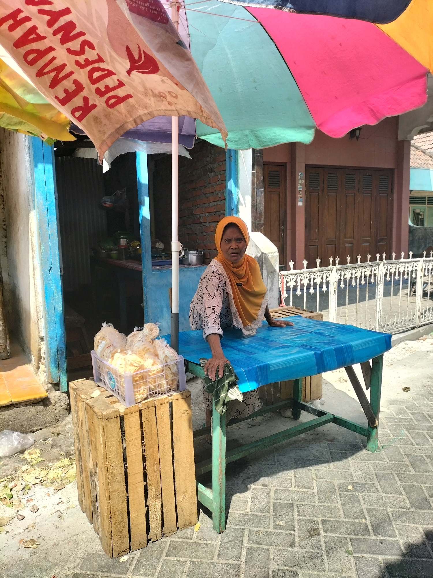 A woman wearing an orange hijab and a patterned shirt stands behind a blue-covered table. She is positioned in front of a stall with a blue doorway, under a multi-colored umbrella. To her left, a wooden crate holds a plastic container filled with packaged snacks. The scene appears to be outdoors, with a paved walkway and a building with wooden doors visible in the background. The umbrella has writing in red on a white banner. The overall environment suggests a street-side market stall. The woman's left hand rests on the table, and her right arm is bent, with her hand partially obscured. The table is green with a blue tablecloth. A white metal fence is in the background. The image is taken in Sembayat, Indonesia.