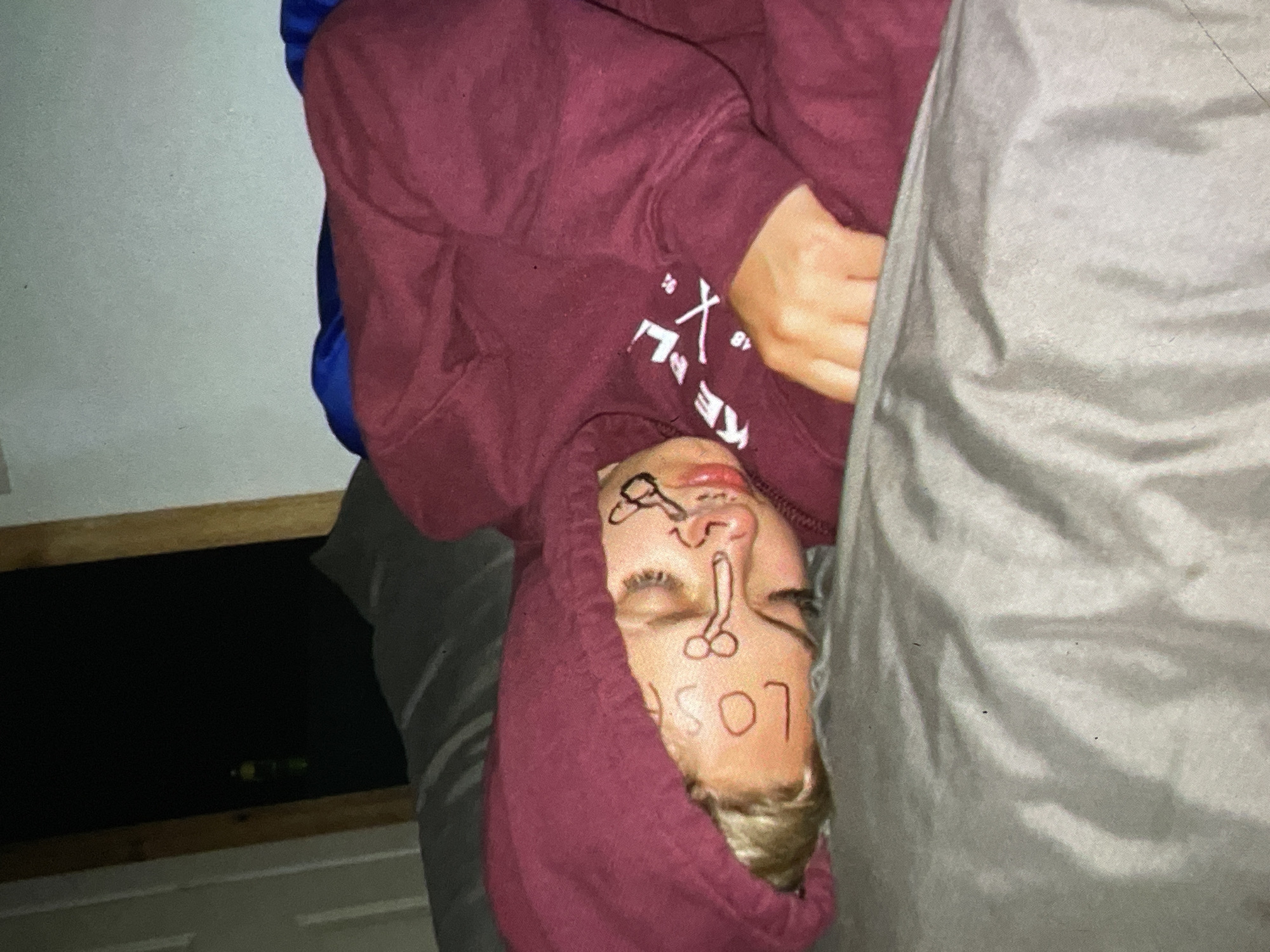 A person is lying on their back, with their head resting on a pillow. They are wearing a maroon hooded sweatshirt, and their face has been drawn on with what appears to be a marker. The drawing includes a phallic symbol, eyes, and the word "LOS." The person's eyes are closed, and their facial expression is unreadable due to the drawing. The scene appears to be indoors, possibly a bedroom, with a grey sheet or blanket covering part of the image. There is a dark object in the background, possibly a shelf or a piece of furniture. The lighting is dim, suggesting it might be nighttime or the room is not well-lit. There are no obvious indications of the weather or the specific time of day beyond the potential for it to be dark. The overall mood is ambiguous, but the drawing on the face suggests a prank or a lighthearted, perhaps drunken, situation. No specific location cues within Harmanli, Bulgaria are visible.