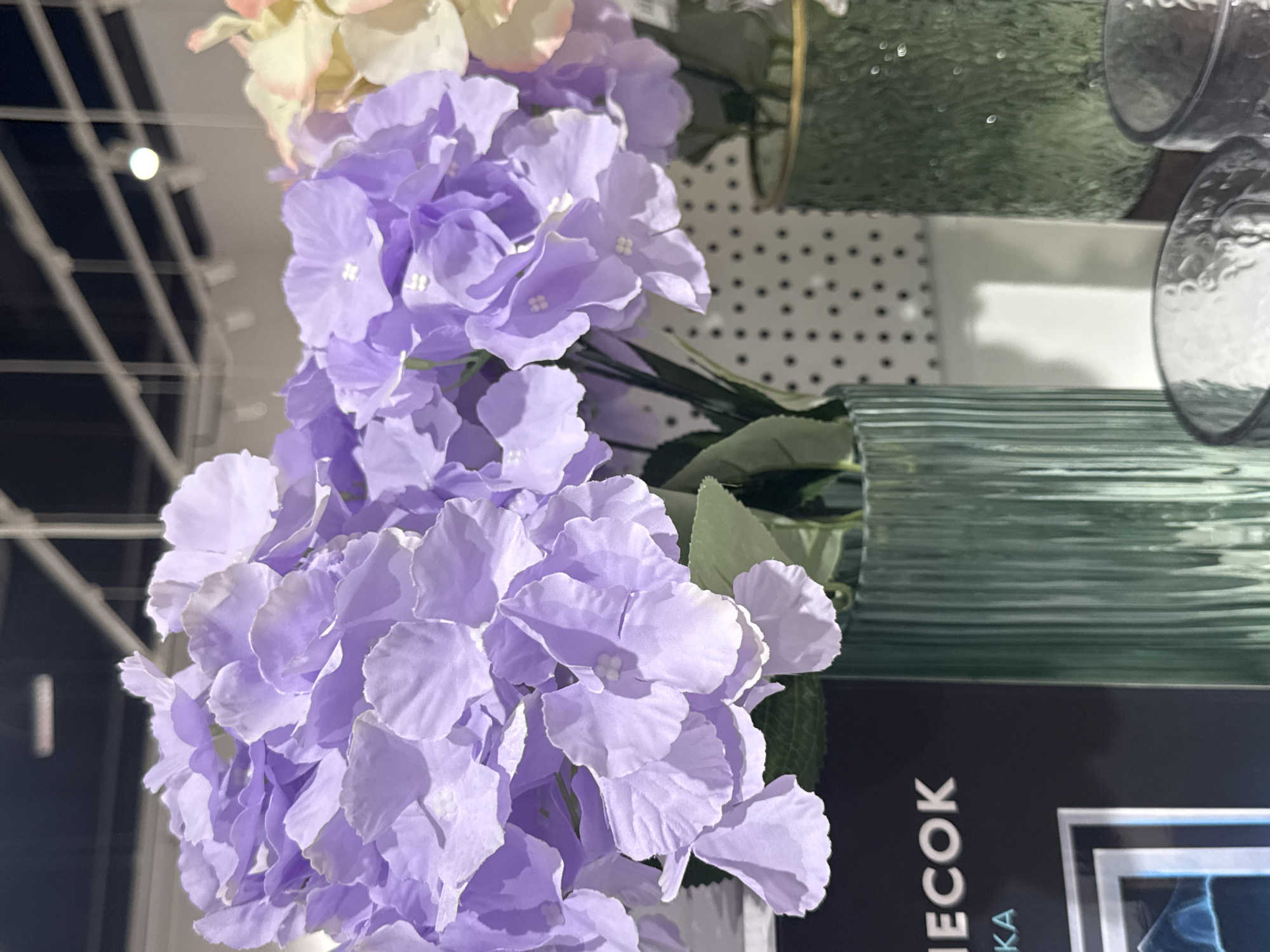 This is a close-up, eye-level shot of artificial purple hydrangeas in a retail environment. The flowers, with their delicate, layered petals, are the primary subject, filling the majority of the frame. They are arranged in a textured, fluted green vase. To the left of the vase, a portion of a dark-colored display with silver text reading "IECOK" is visible. The background features a white surface with a repeating pattern of black dots, and overhead lighting fixtures are visible. The scene appears to be an indoor display, possibly in a home decor store or a flower shop. There is no indication of the city of Ufa, Russia within the image itself.