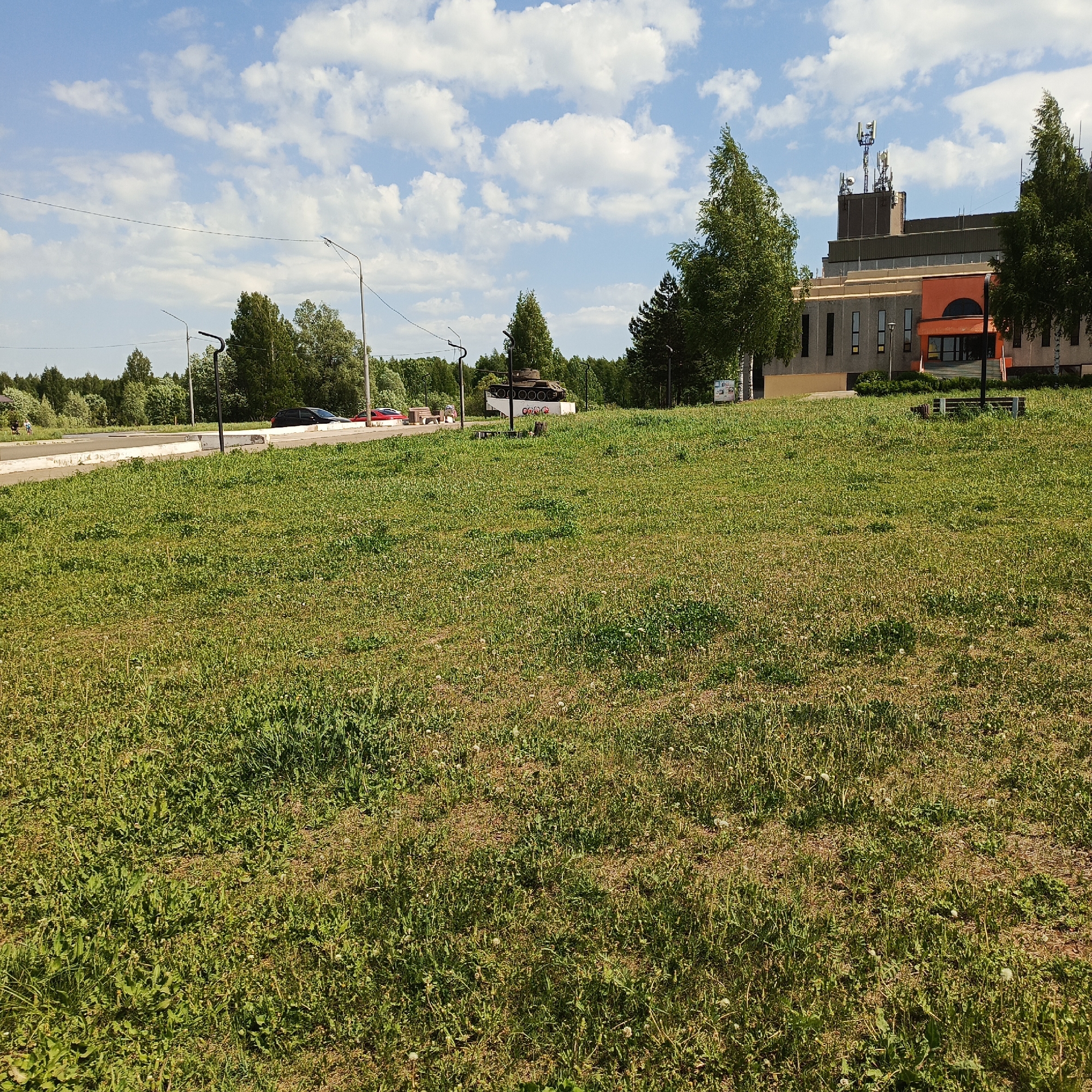 The image depicts a grassy field in Марковский, Russia, with a road and sparse trees in the mid-ground. A building with a modern design, including cell towers on its roof, is located to the right of the field. A static tank memorial is visible along the roadside in the background. A few cars and people are also visible on the side of the road. The sky is blue with scattered clouds, suggesting daytime and fair weather.
