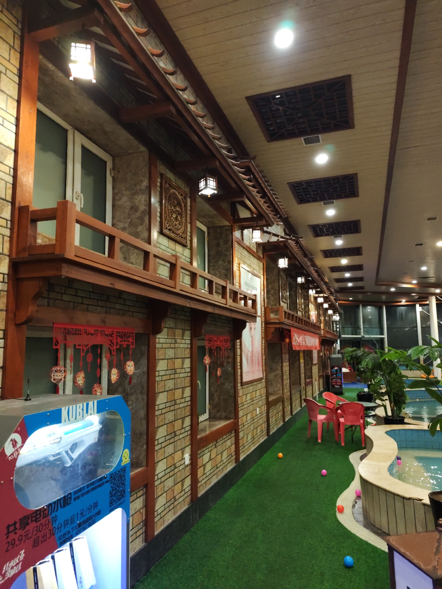 The image depicts an indoor recreational facility with a distinct East Asian architectural influence, located in Lindian Town, China. The scene features a multi-level structure with wooden balconies and eaves, reminiscent of traditional Chinese buildings. The walls are constructed with patterned tiles, and red paper decorations hang from some of the balconies.
The floor is covered with artificial turf, giving the space a garden-like feel. Scattered across the turf are several colorful plastic balls, suggesting a play area. Adjacent to the turf is a swimming pool with a curved, light-colored edge. A few red chairs are placed near the pool.
In the foreground, a prominent blue and red machine with a clear plastic display case shows a white toy gun inside. Text in Chinese is visible on the machine, indicating it may be a rental or vending machine for the toy. Green plants are situated near the pool, adding to the relaxed, leisure atmosphere. The ceiling is made of wooden planks with recessed square decorative panels and illuminated with ceiling lights. The overall impression is that of a family-friendly entertainment or relaxation center. There are no people clearly visible in the scene.