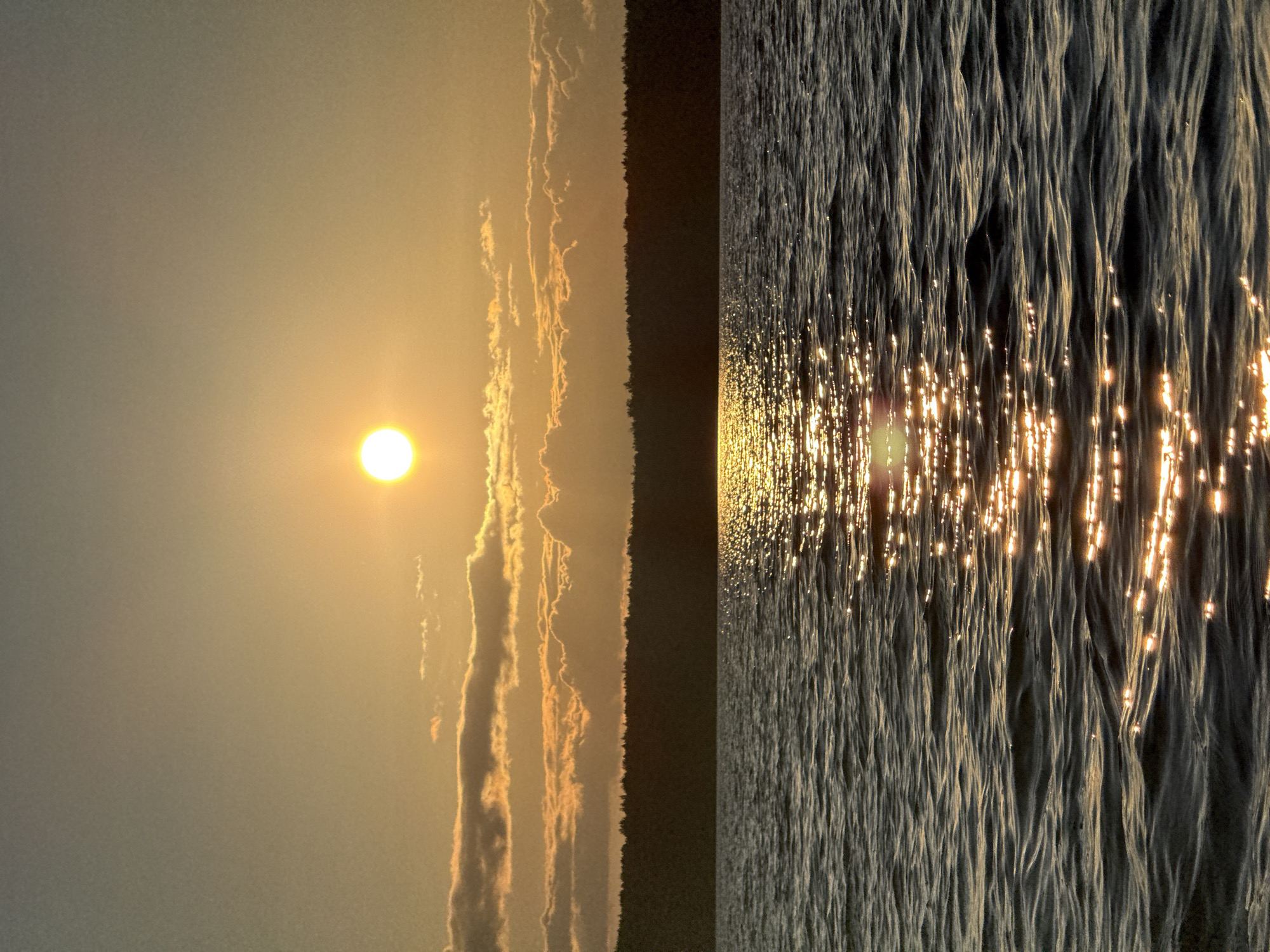 The image depicts a serene sunset over a body of water. A bright, round sun is visible in the upper center of the frame, casting a warm, golden glow across the sky. Wispy clouds, tinged with orange and yellow, are scattered across the horizon. Below the sky, a dark, silhouetted tree line forms a distinct border, suggesting a wooded area or forest. The foreground is dominated by the rippling surface of a lake or sea, reflecting the sunlight in a shimmering pathway. The water is dark, with gentle waves creating texture and movement. The overall atmosphere is tranquil and natural, with no signs of human presence or activity. The location in Gora (Khokhlovskoe S/P), Russia, is implied by the provided information.