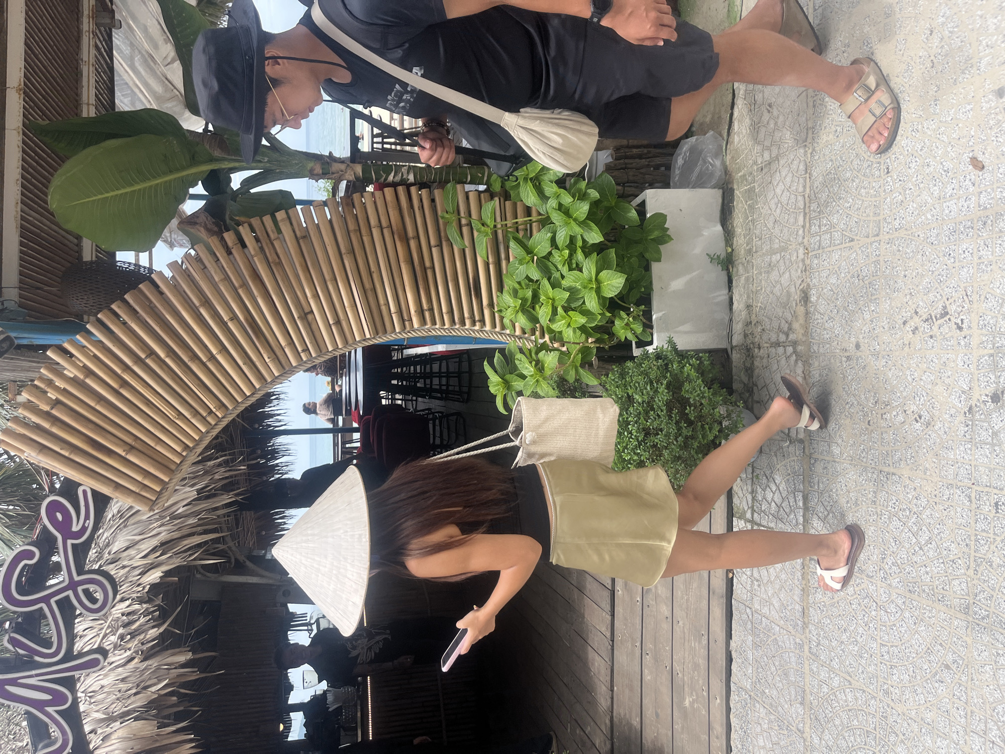 The image depicts a street scene in Đà Nẵng, Vietnam. A woman, facing away from the camera, walks on a tiled walkway. She is wearing a conical hat, a black tank top, a satin skirt, and white sandals. She holds a pink smartphone in her left hand. A woven bag hangs from her right shoulder. To her right, a man walks in the same direction. He is wearing a black t-shirt, black shorts, a black hat, and sandals. He carries a bag slung across his body.
In the background, a thatched-roof structure resembling a cafe or restaurant is visible, with a curved bamboo archway leading towards an outdoor seating area. Green plants in a white planter are positioned near the bamboo arch. The ocean and a distant view of a beach can be seen behind the structure. The overall setting suggests a casual, tropical environment.