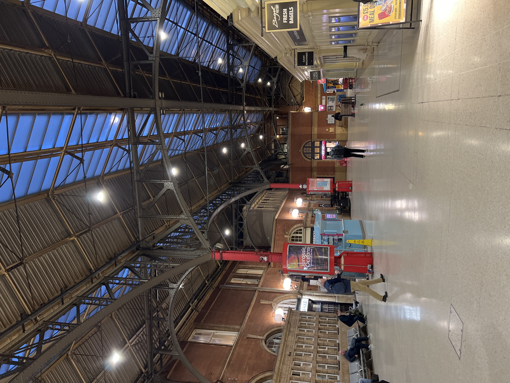 The image depicts the interior of a train station with a high, arched glass and metal roof. The floor is polished and reflective, showing the overhead lighting and structure. Several people are visible in the station, some sitting on benches, others walking. There are various kiosks and shops lining the sides of the station, including a "Bagel Factory" and a kiosk advertising "Jurassic Oceans." The overall atmosphere is that of a busy public transport hub.